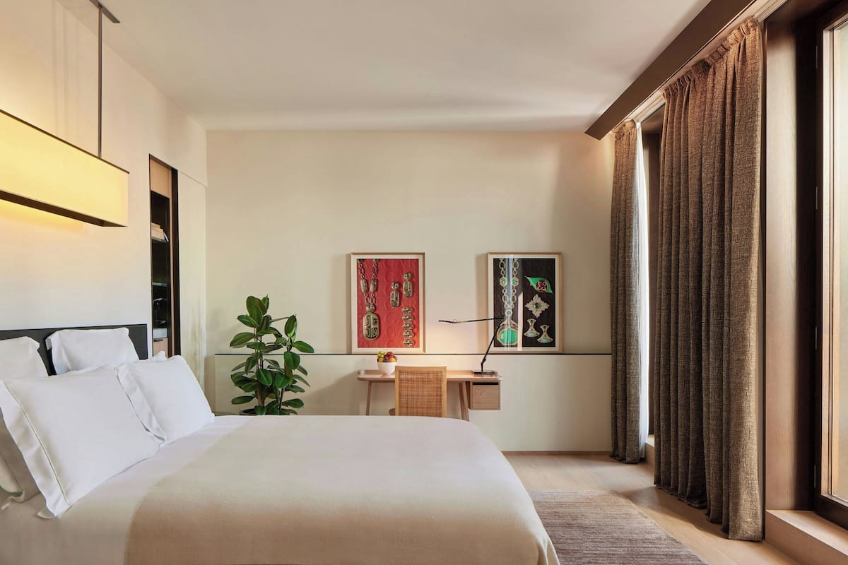 A hotel room with a bed, a desk, a plant, and artwork on the wall.