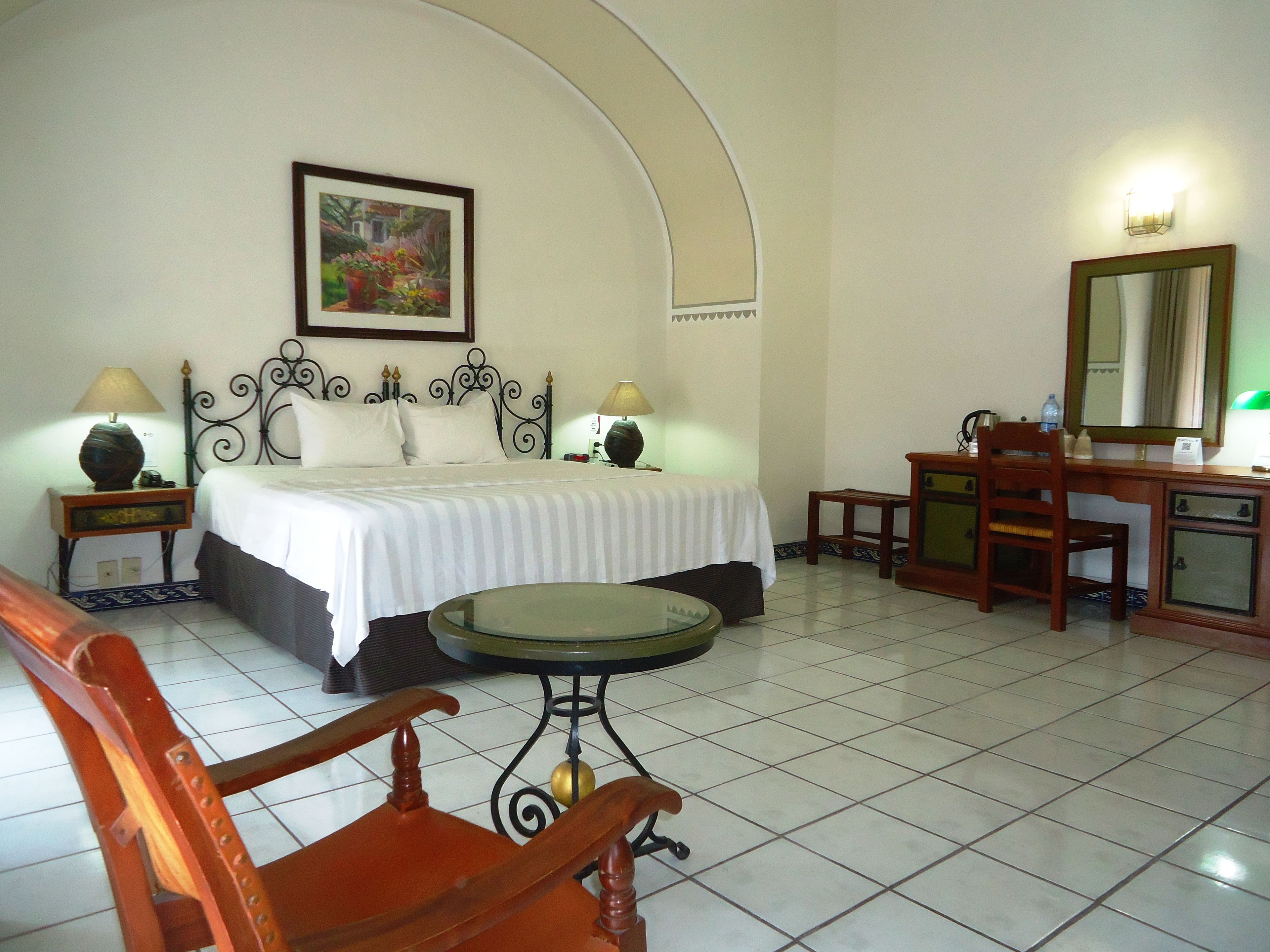 A hotel room with a bed, a desk, a chair, a table, and a painting on the wall.