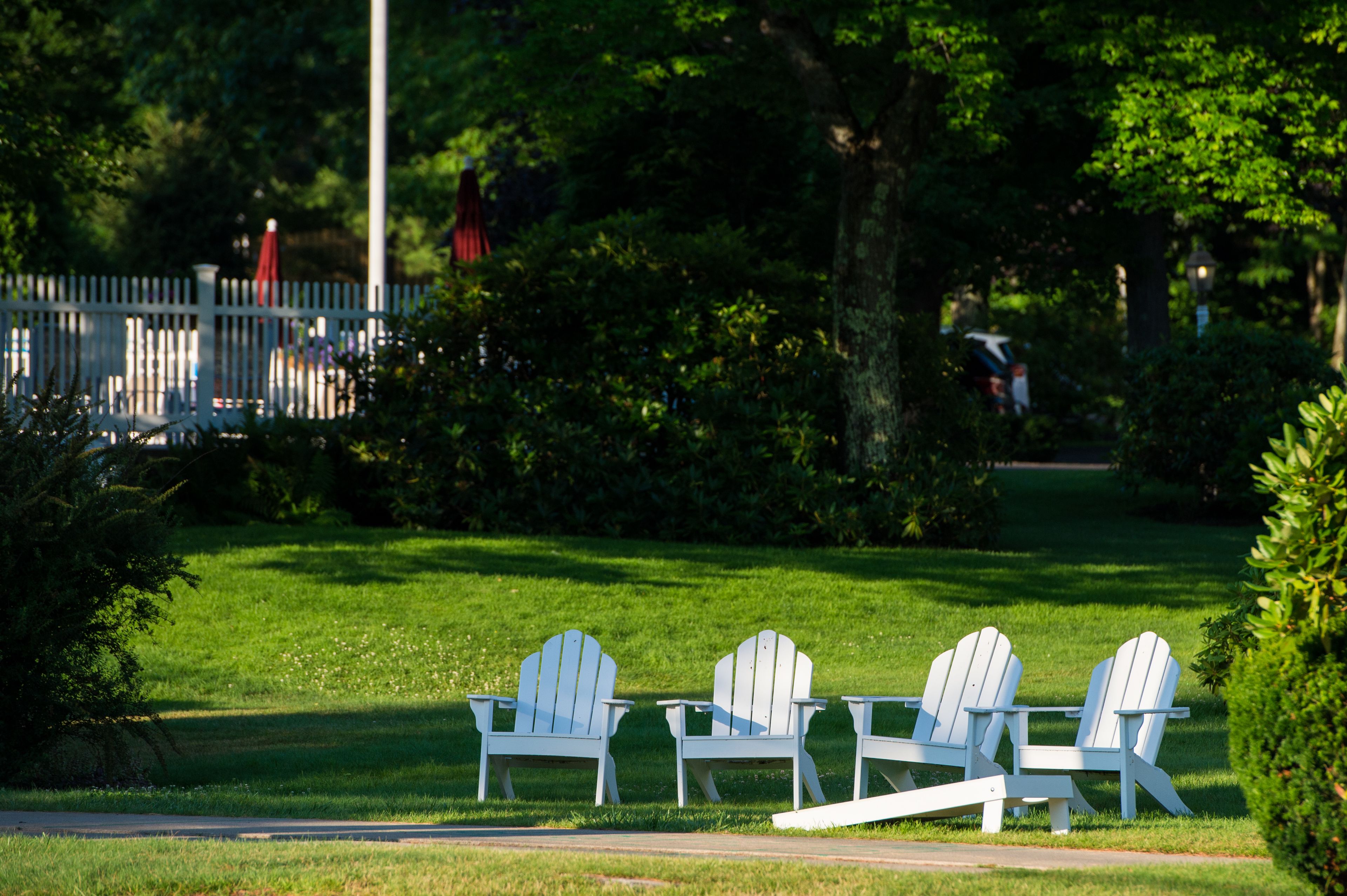 Three white Adirondack chairs on a grassy lawn with a white picket fence in the background.