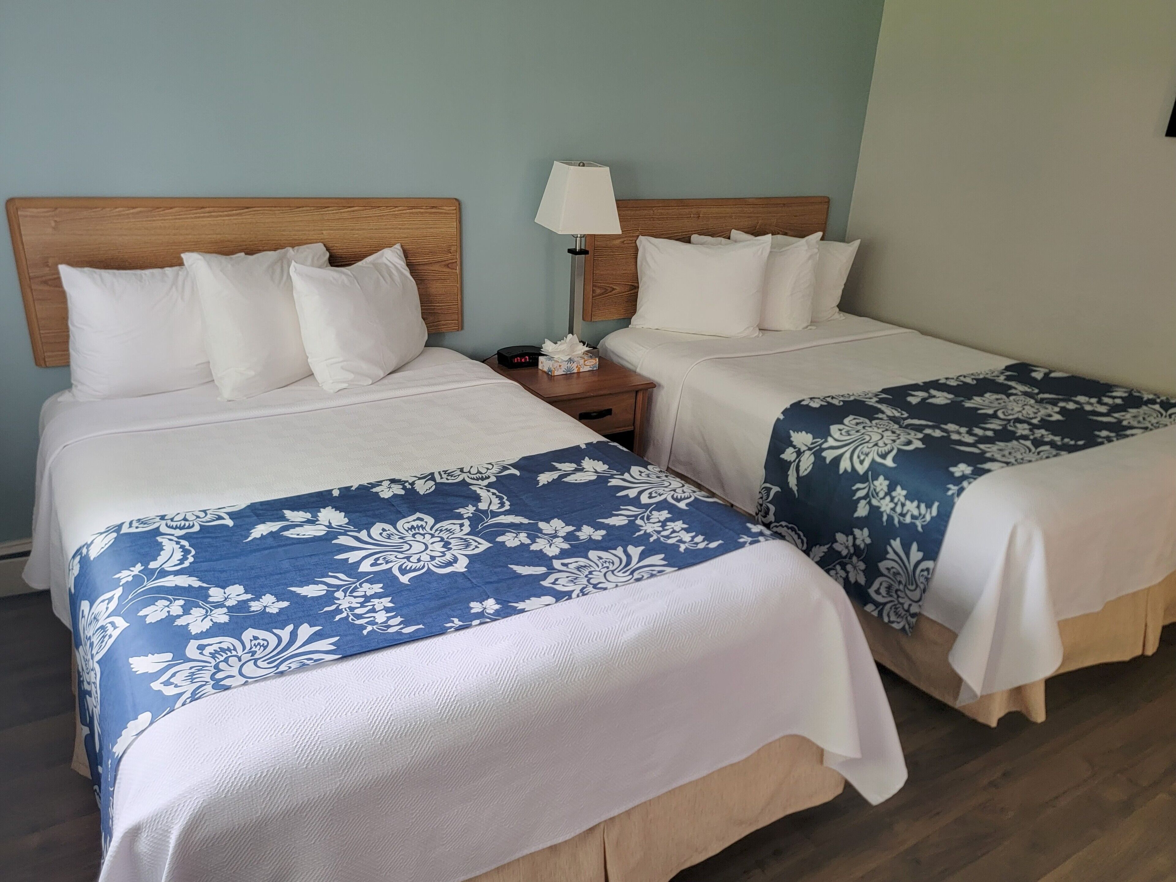 Two double beds with wooden headboards, white bedding, and blue floral bedspreads in a room with light blue walls and a wooden floor.
