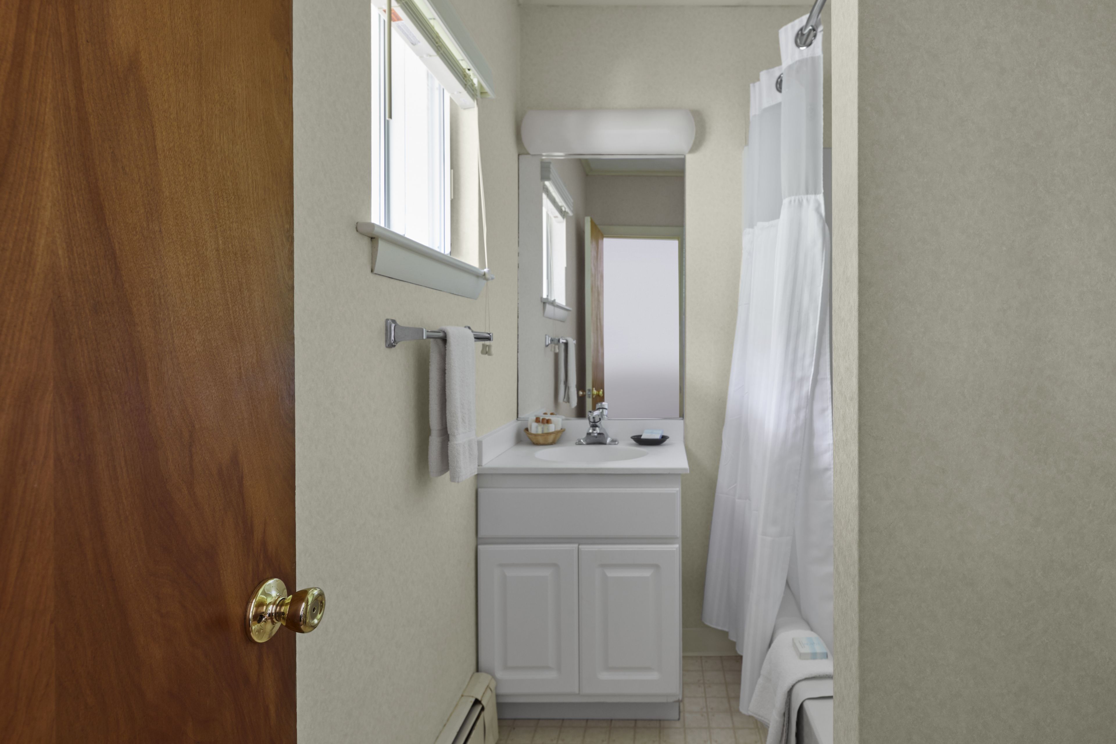 A bathroom with a wooden door, white vanity, mirror, and white shower curtain.