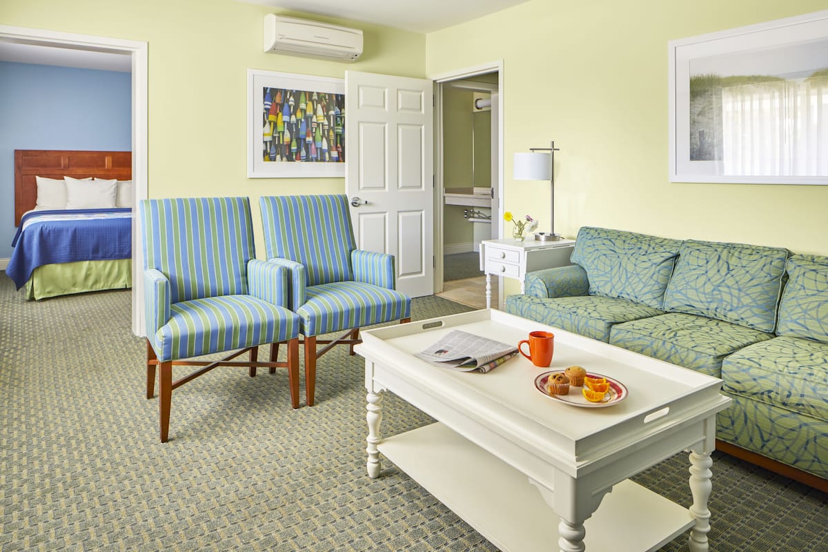 Une chambre d’hôtel avec un lit, deux fauteuils à rayures, un canapé, une table basse blanche sur laquelle se trouve un plateau de muffins, et un tableau accroché au mur.