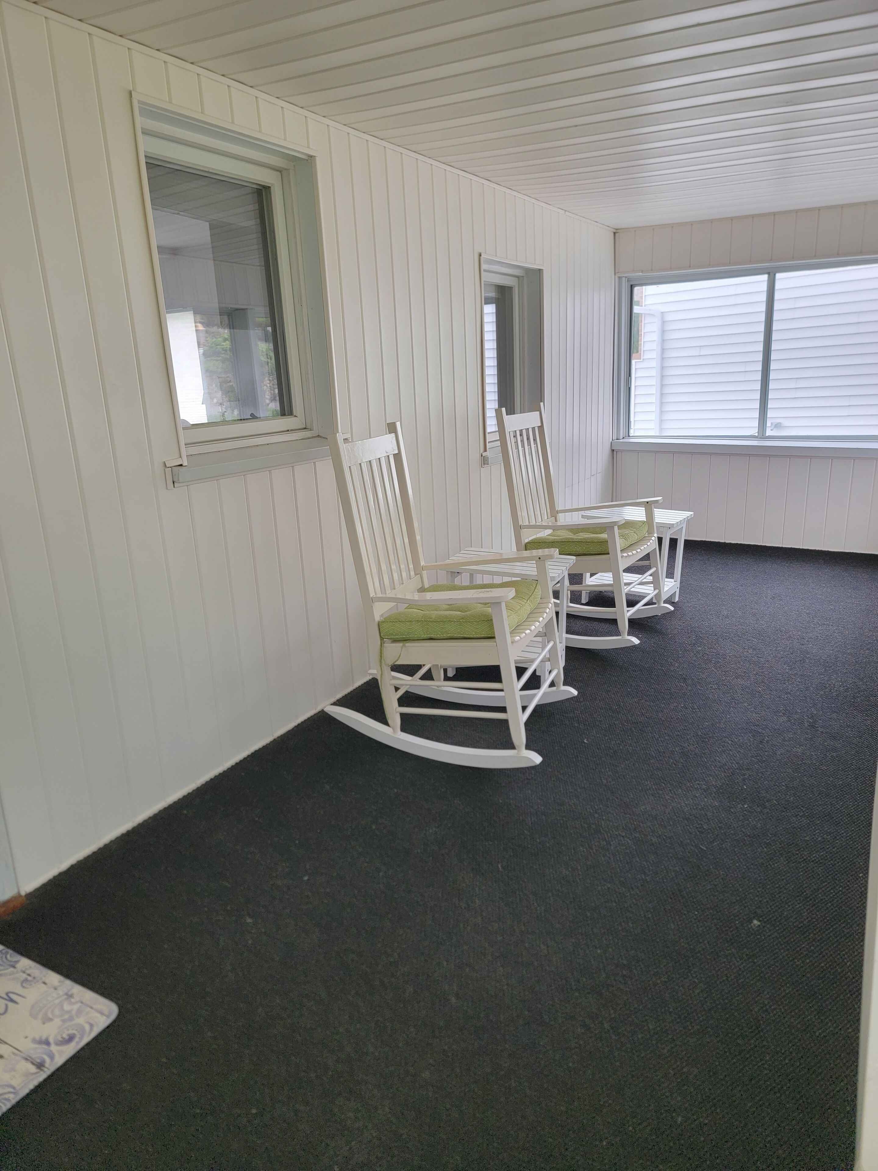 A room with white paneled walls, a black carpet, and three rocking chairs with green cushions.