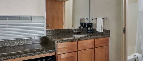 A bathroom with a granite countertop, wooden cabinets, a sink, a coffee maker, and a shower curtain.