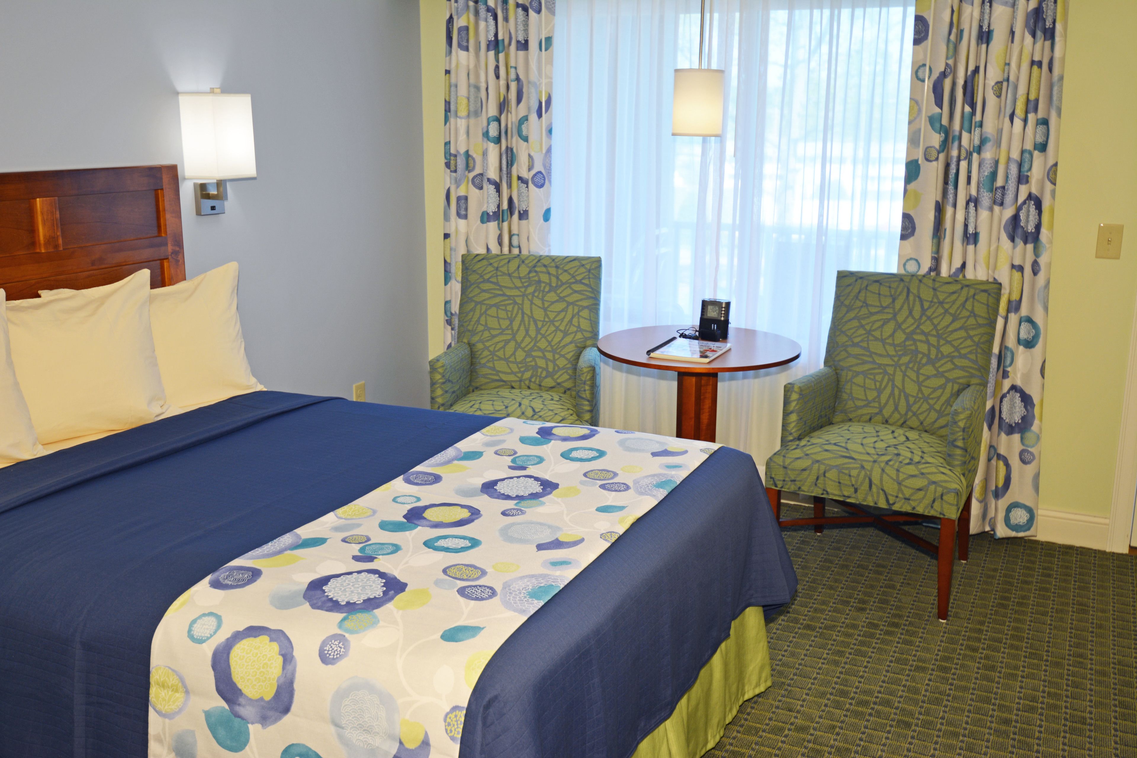 A hotel room with a bed, two chairs, a small table, and a lamp.