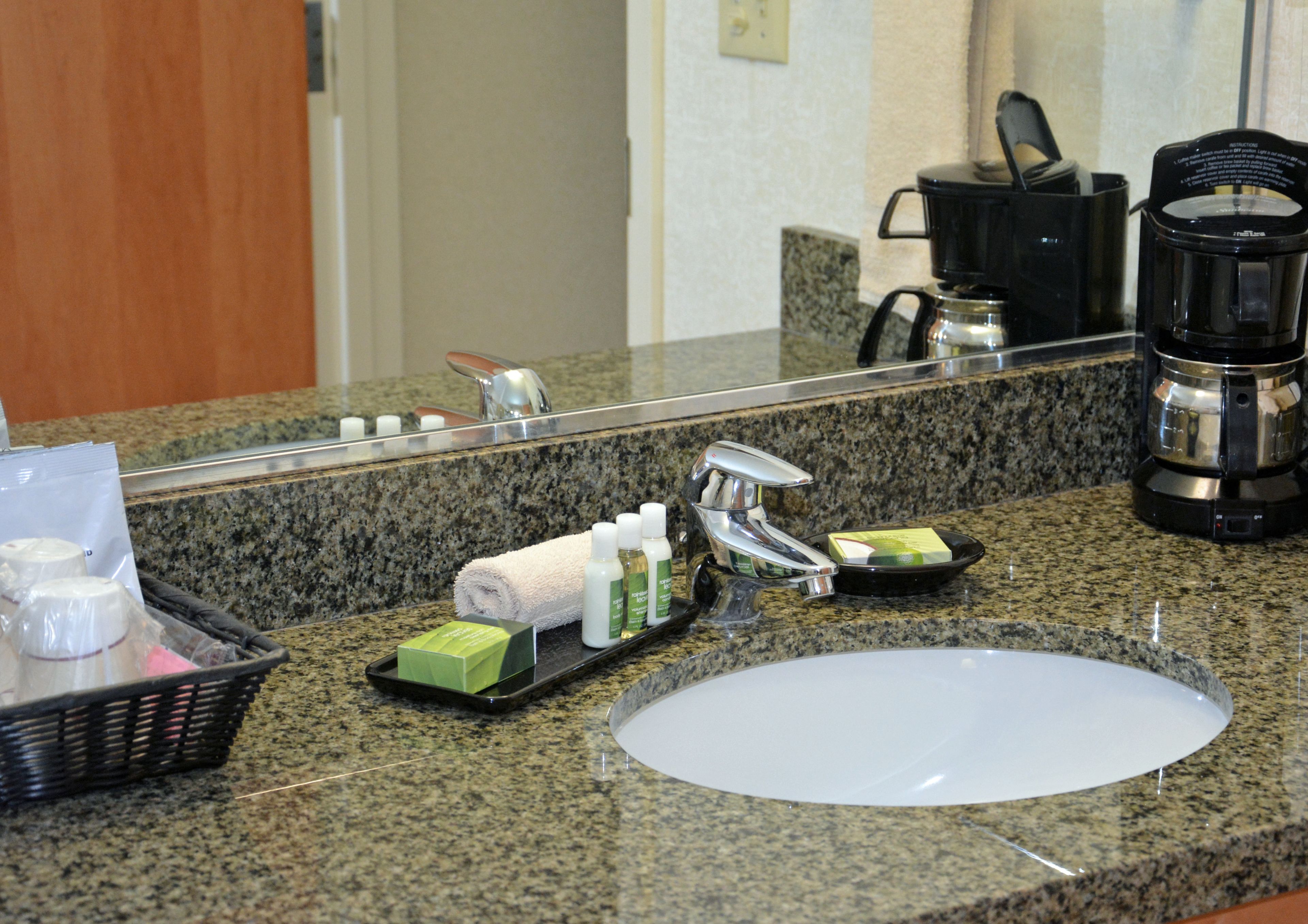 A hotel bathroom with a granite countertop, a sink, a mirror, a coffee maker, and toiletries.