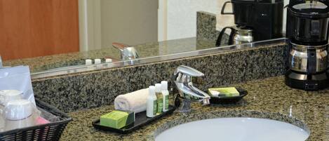 A hotel bathroom with a granite countertop, a sink, a mirror, a coffee maker, and toiletries.