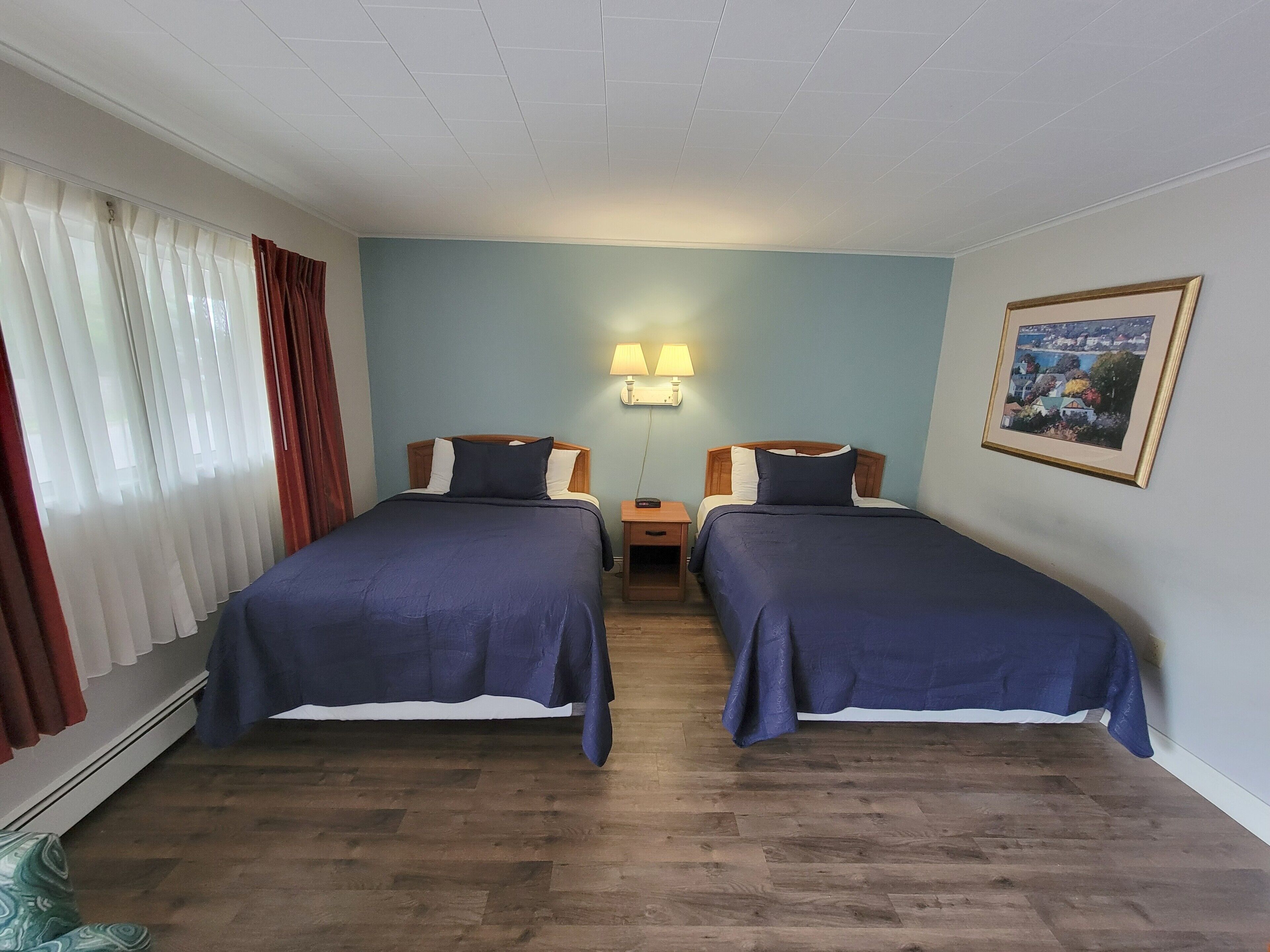 A hotel room with two beds, wooden flooring, a nightstand with a lamp, and a framed picture on the wall.
