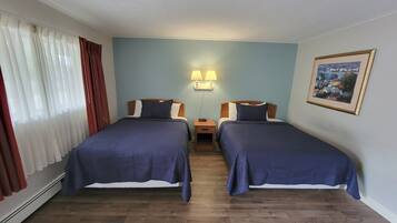 A hotel room with two beds, wooden flooring, a nightstand with a lamp, and a framed picture on the wall.