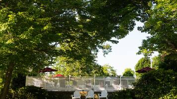 A fenced outdoor seating area with white chairs and red umbrellas, surrounded by lush greenery and trees.