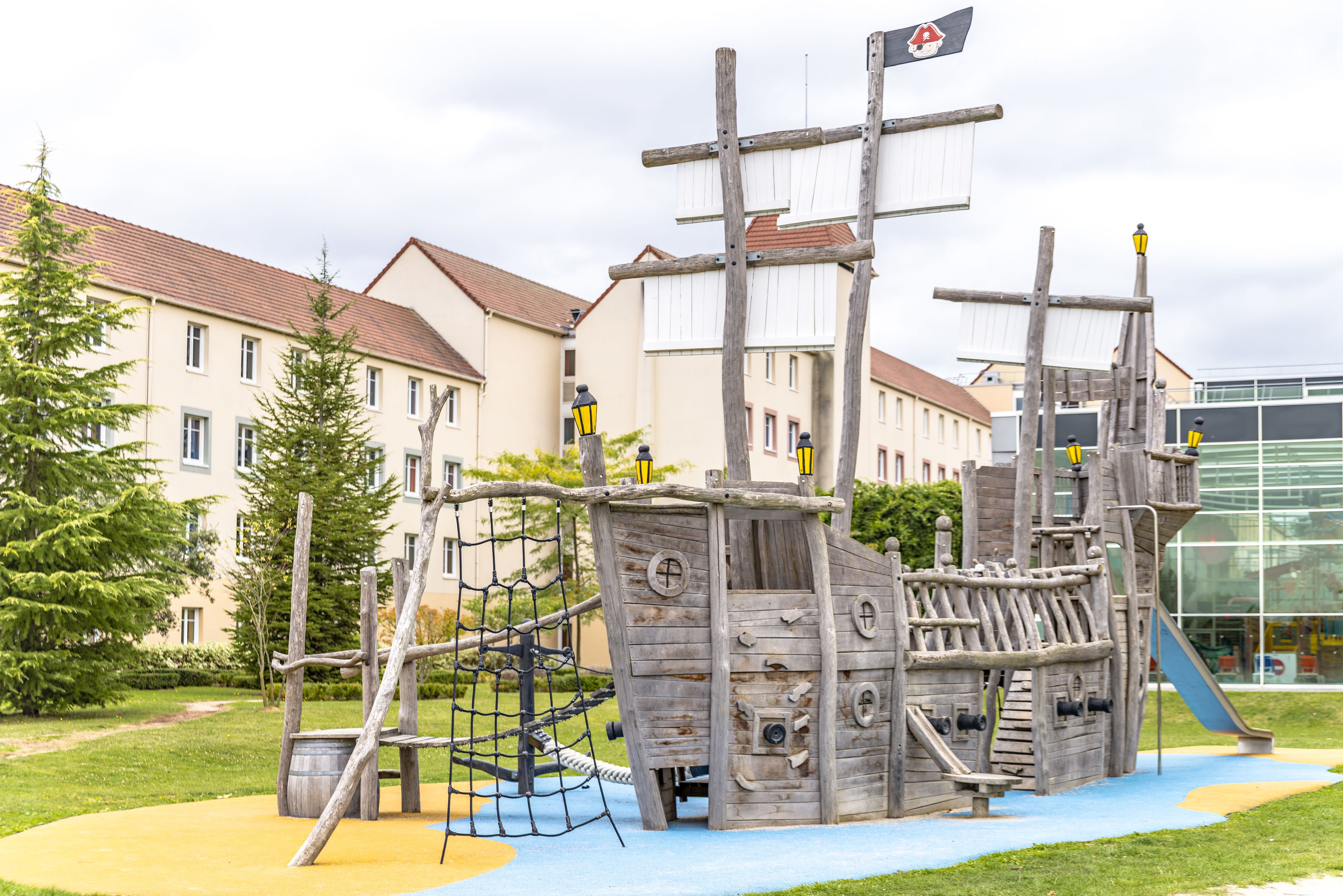 Una estructura de juego de barco pirata de madera en un parque, con un edificio al fondo.