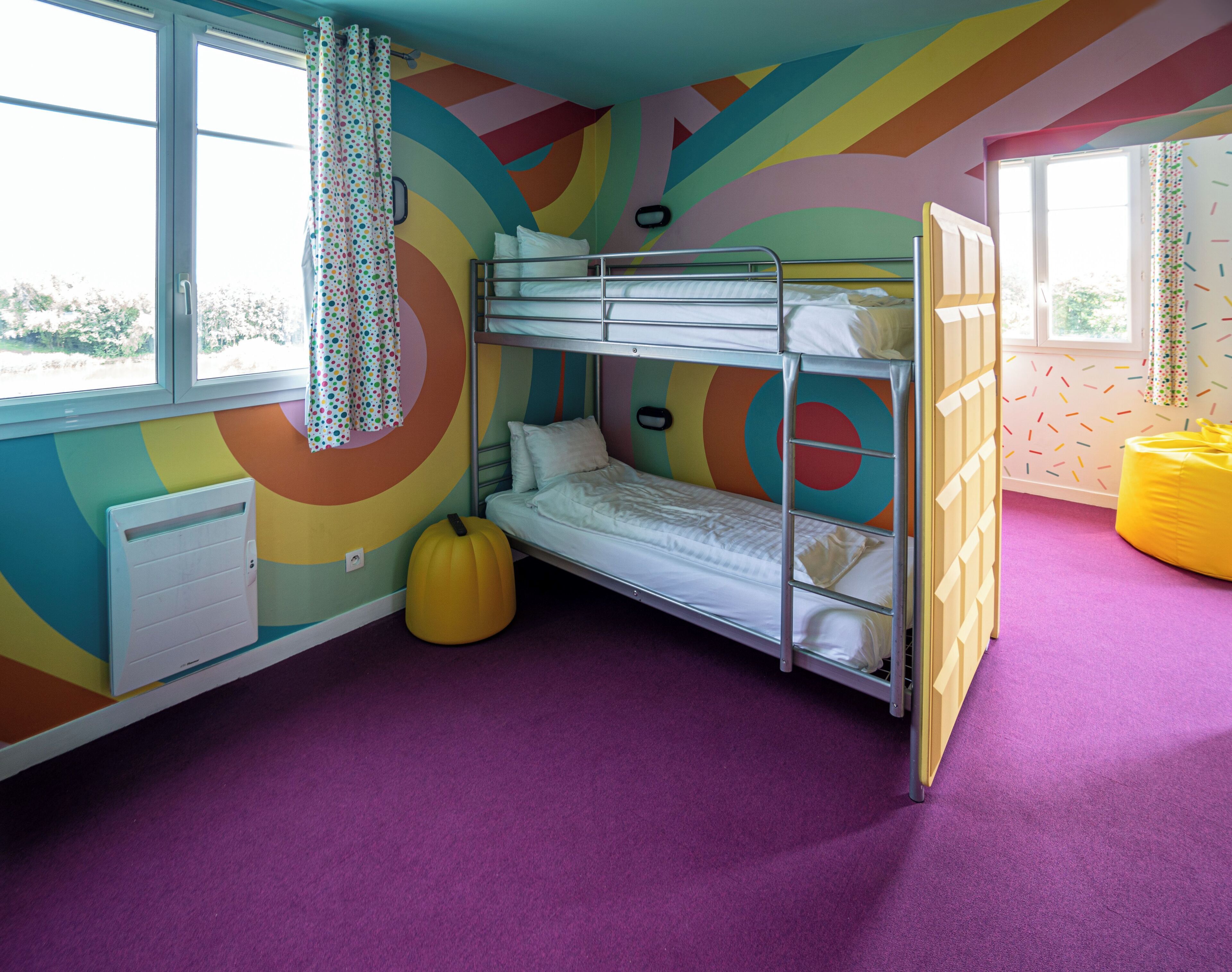 Une chambre avec un lit superposé, un mur aux motifs colorés, une fenêtre avec des rideaux, un radiateur et un fauteuil en forme de sac de haricots.