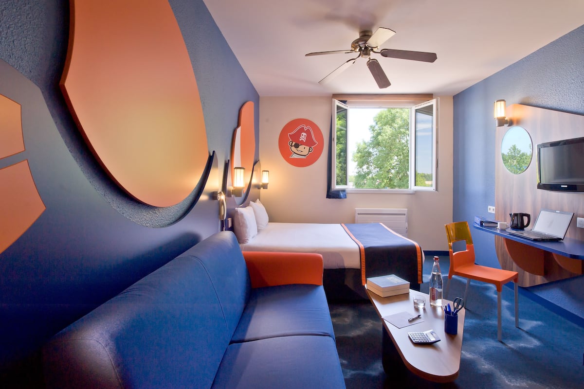 A modern hotel room with a bed, a sofa, a desk with a laptop, a chair, and a TV.