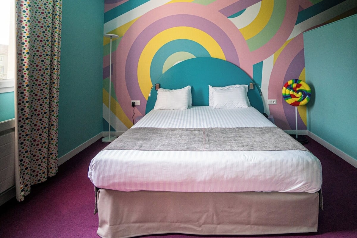 A bed with white bedding, a colorful abstract wall mural, and a lollipop-shaped decoration.