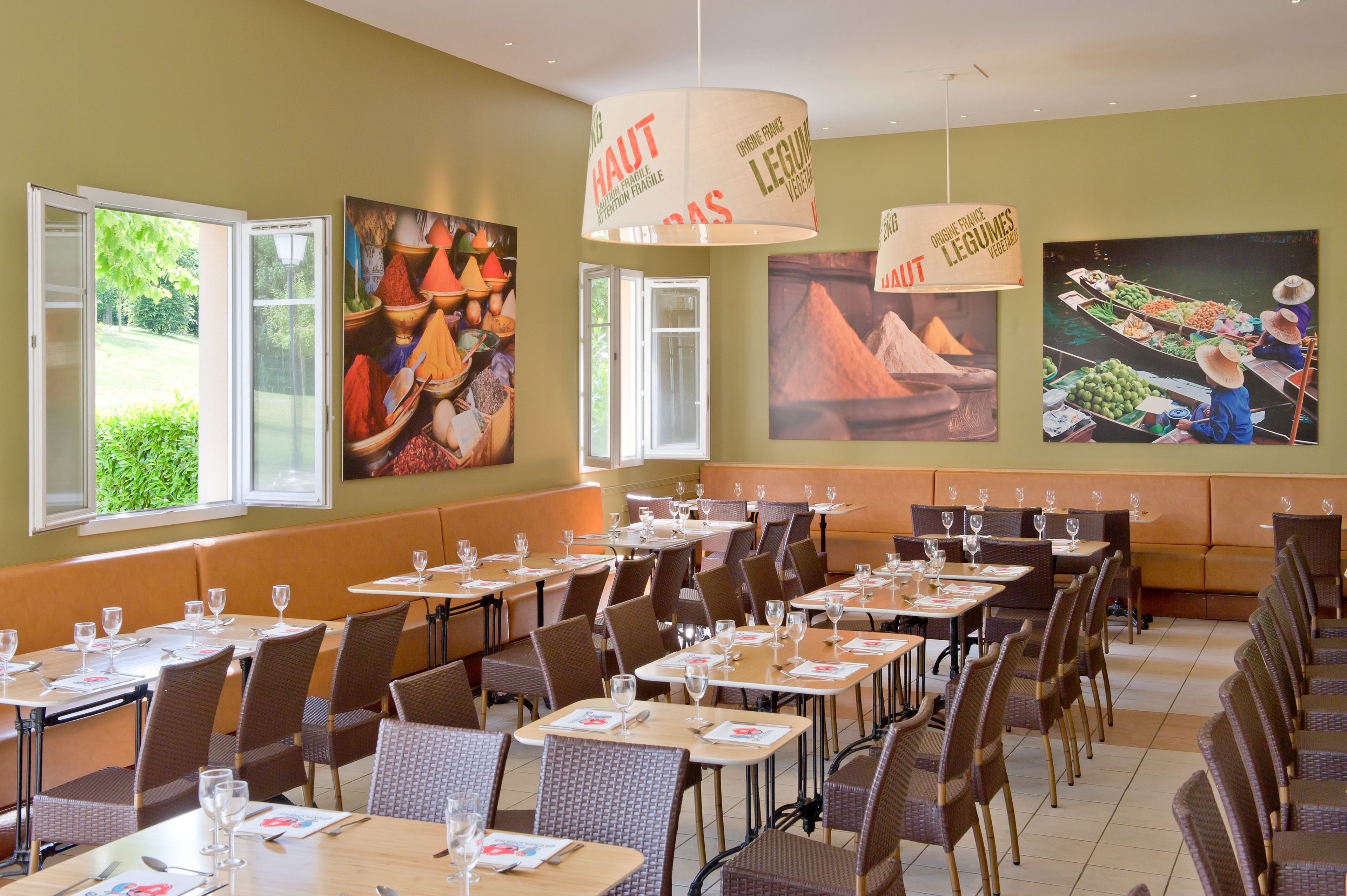 A restaurant with tables set for dining, large paintings on the walls, and a view of greenery outside.