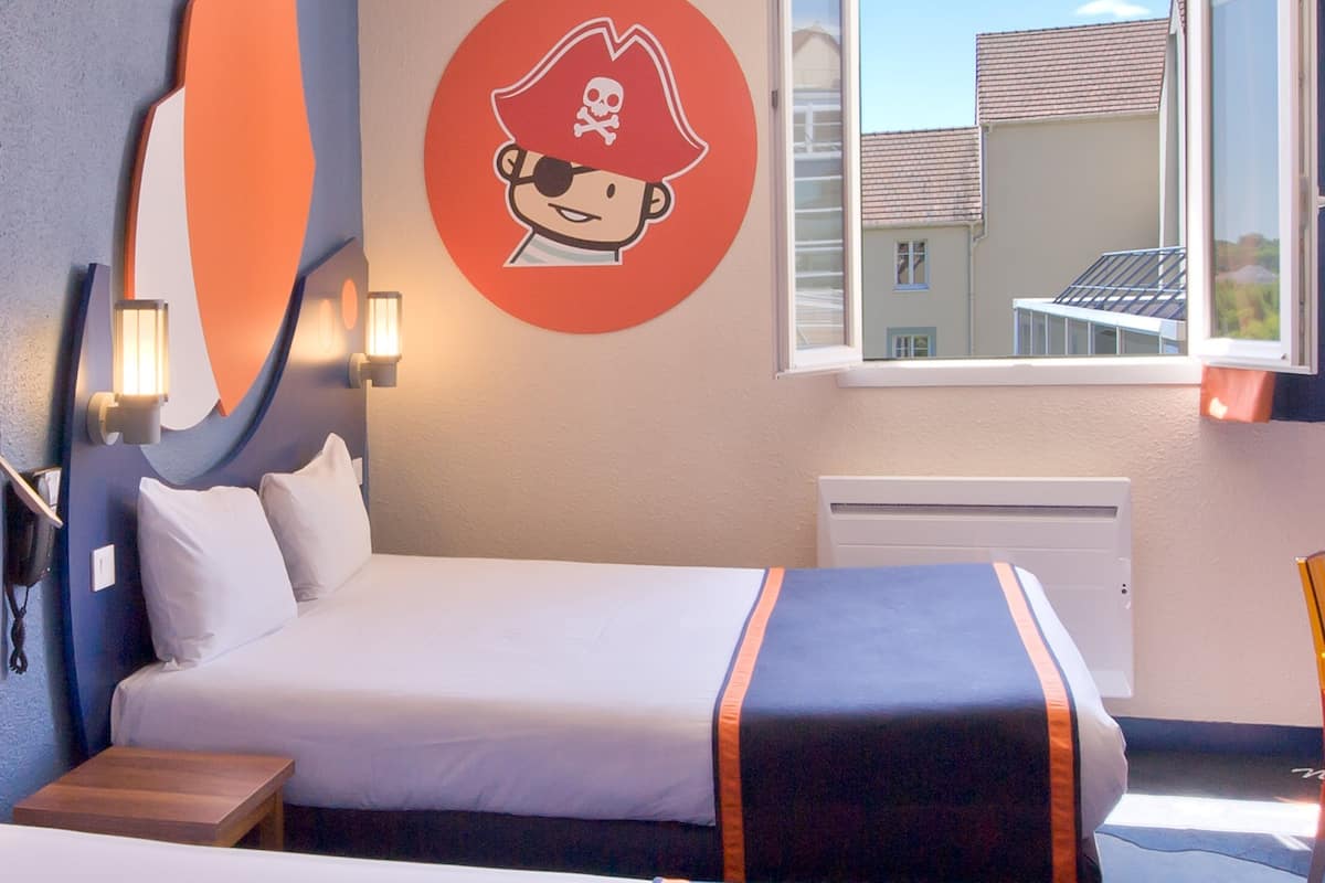 A hotel room with a bed, a wall decoration of a pirate, and a window with a view of buildings.