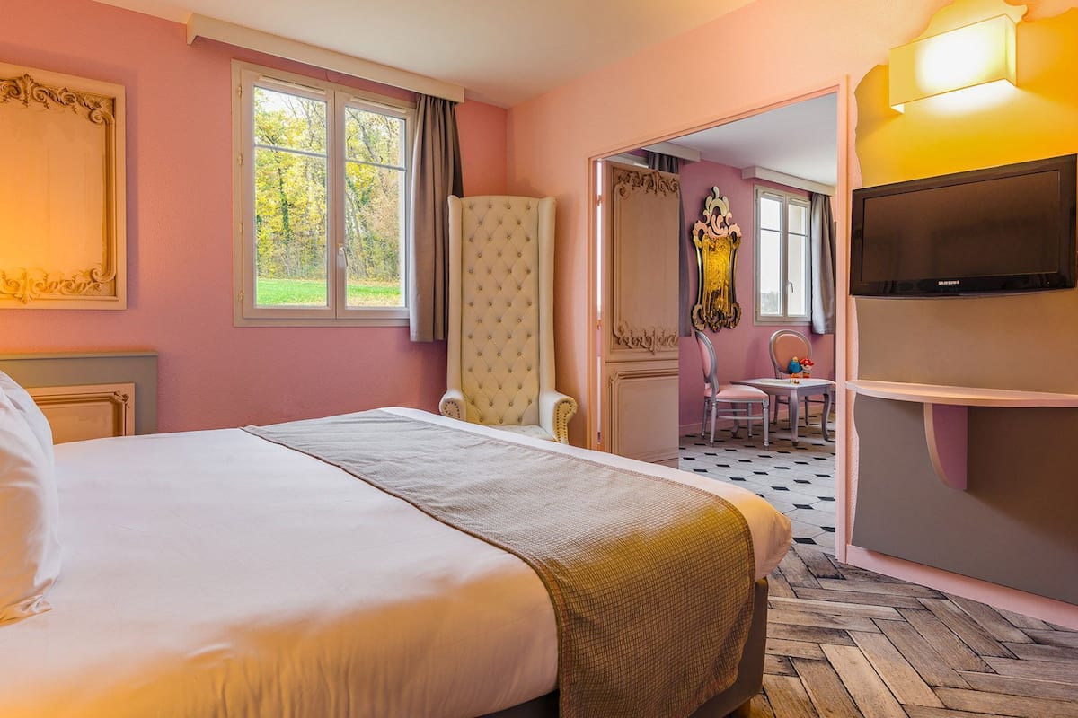 A hotel room with a large bed, a television, a window with curtains, and a mirror.