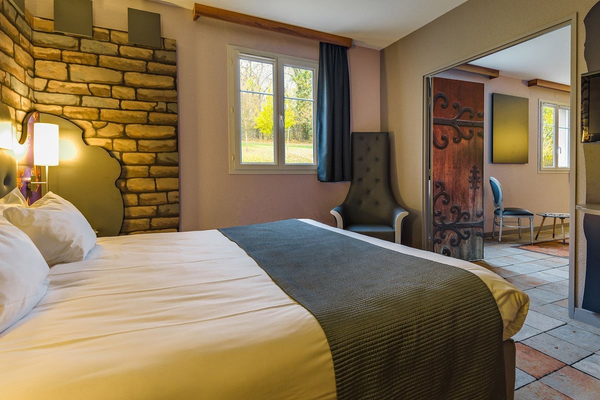 A hotel room with a stone accent wall, a bed with white linens, a chair, a desk, and a television.