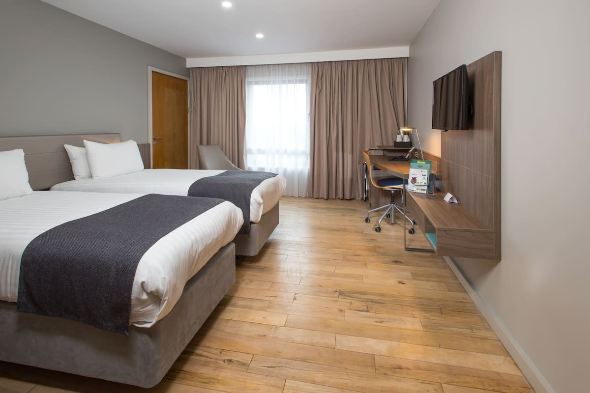 Ein Hotelzimmer mit zwei Betten, einem Schreibtisch mit Stuhl, einem Fernseher und einem großen Fenster mit Vorhängen.