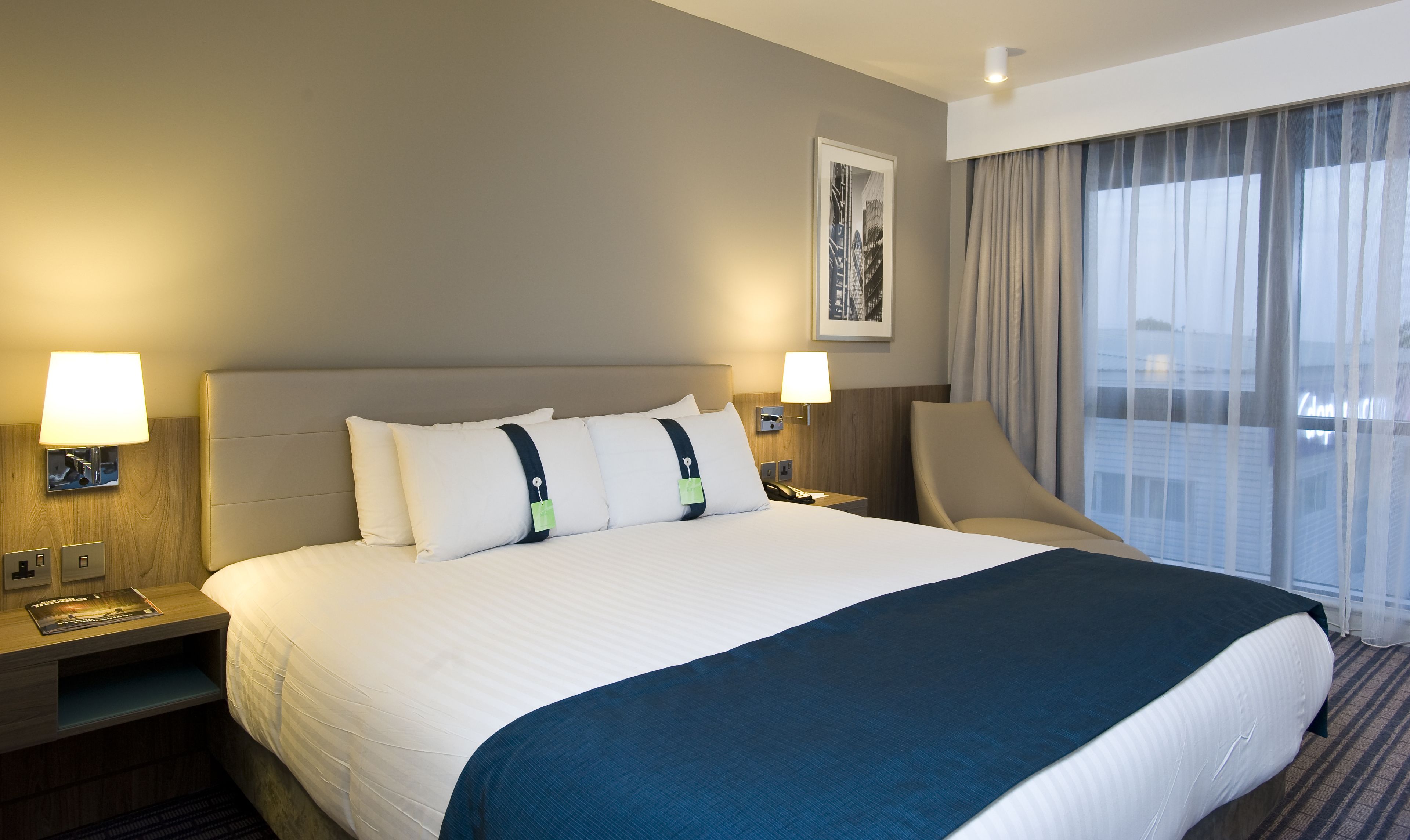 Una habitación de hotel con una cama grande, dos lámparas de noche, una silla, una ventana con cortinas y un cuadro en la pared.