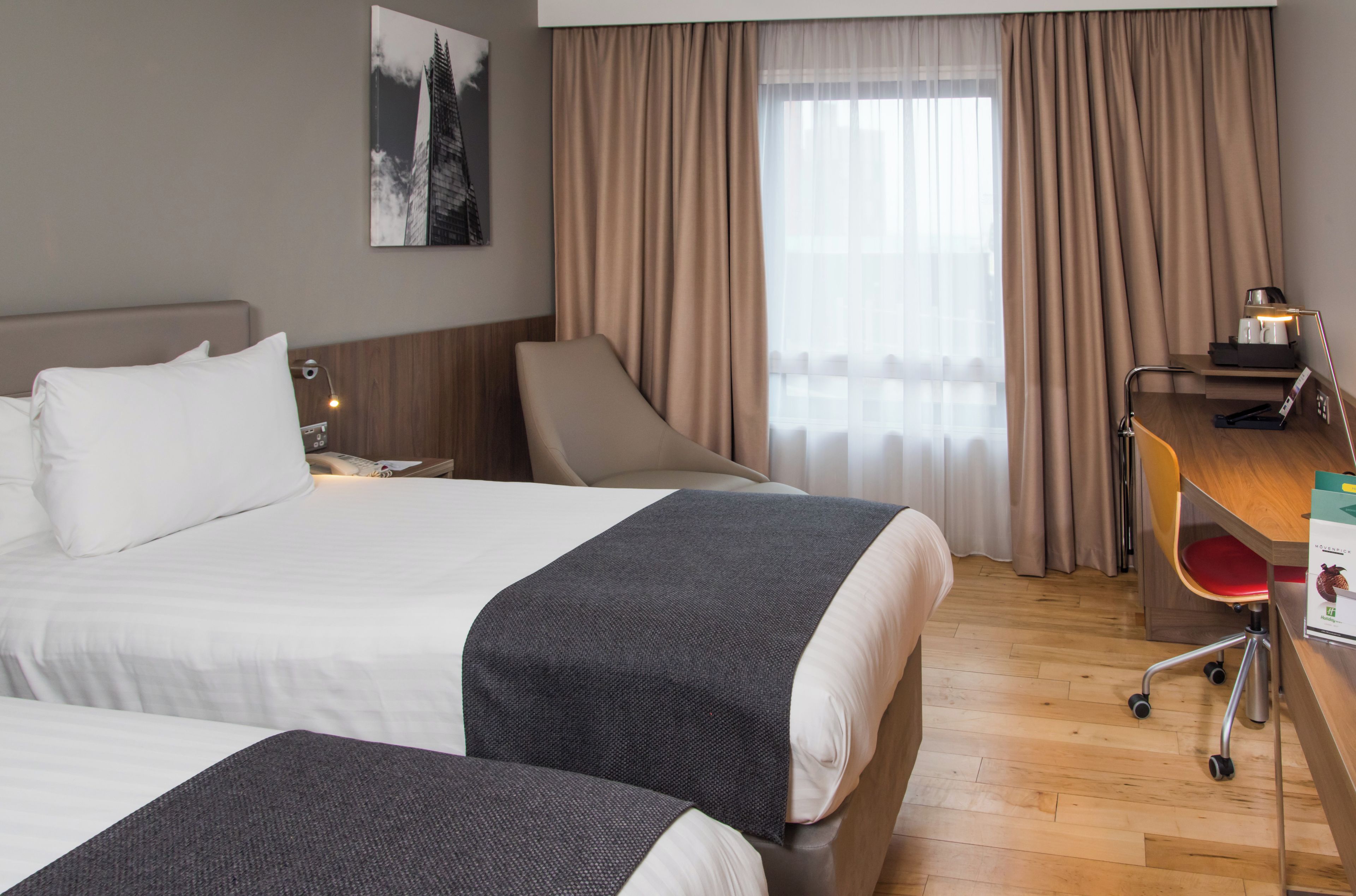 A hotel room with two beds, a desk with a chair, a lamp, and a window with curtains.