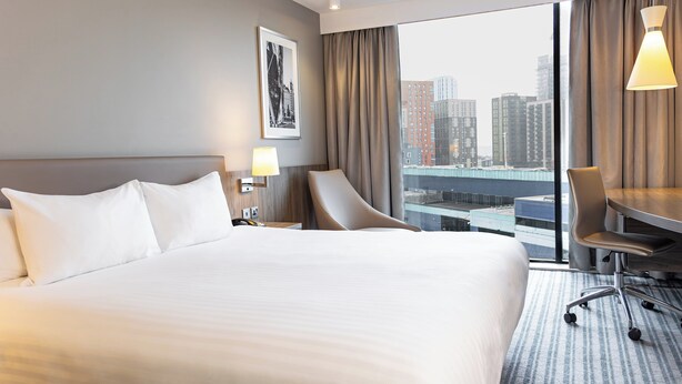 A hotel room with a large bed, a desk, a chair, and a view of a cityscape through the window. - Holiday Inn London - West by IHG (London)