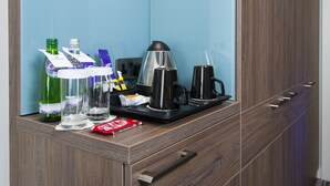 A hotel room with a wooden wardrobe, a blue wall, and a small table with a coffee maker, glasses, and bottles. - Holiday Inn London - West by IHG (London)