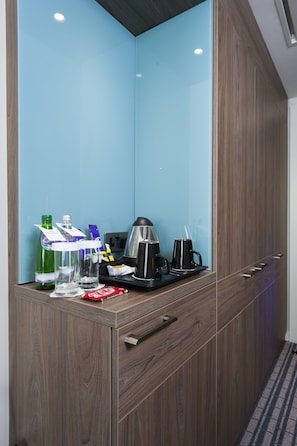 Een hotelkamer met een houten garderobe, een blauwe wand en een klein tafeltje met een koffiezetapparaat, glazen en flessen.