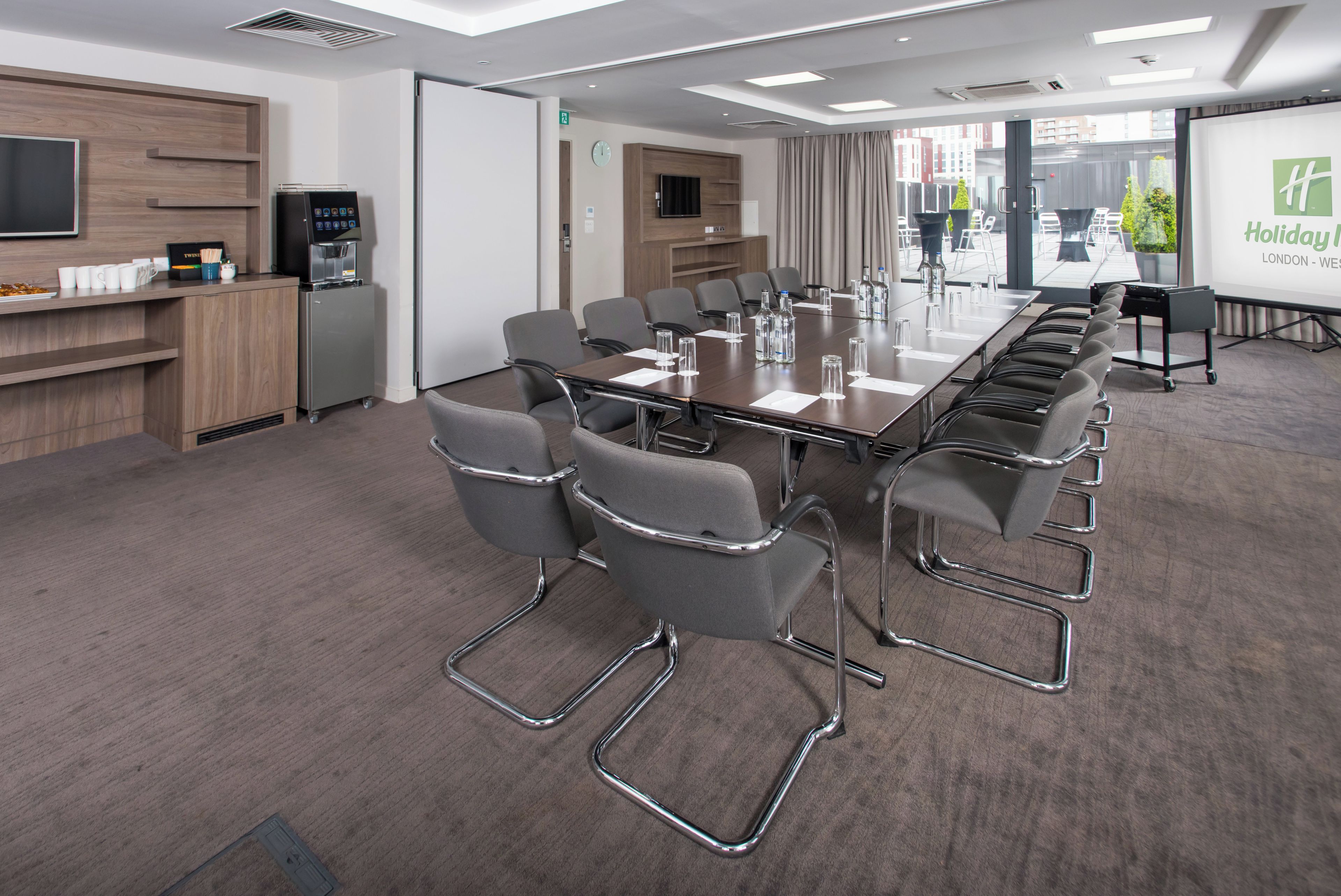 A conference room with a long table, grey chairs, a coffee station, and a projection screen displaying 'Holiday Inn London - West'.