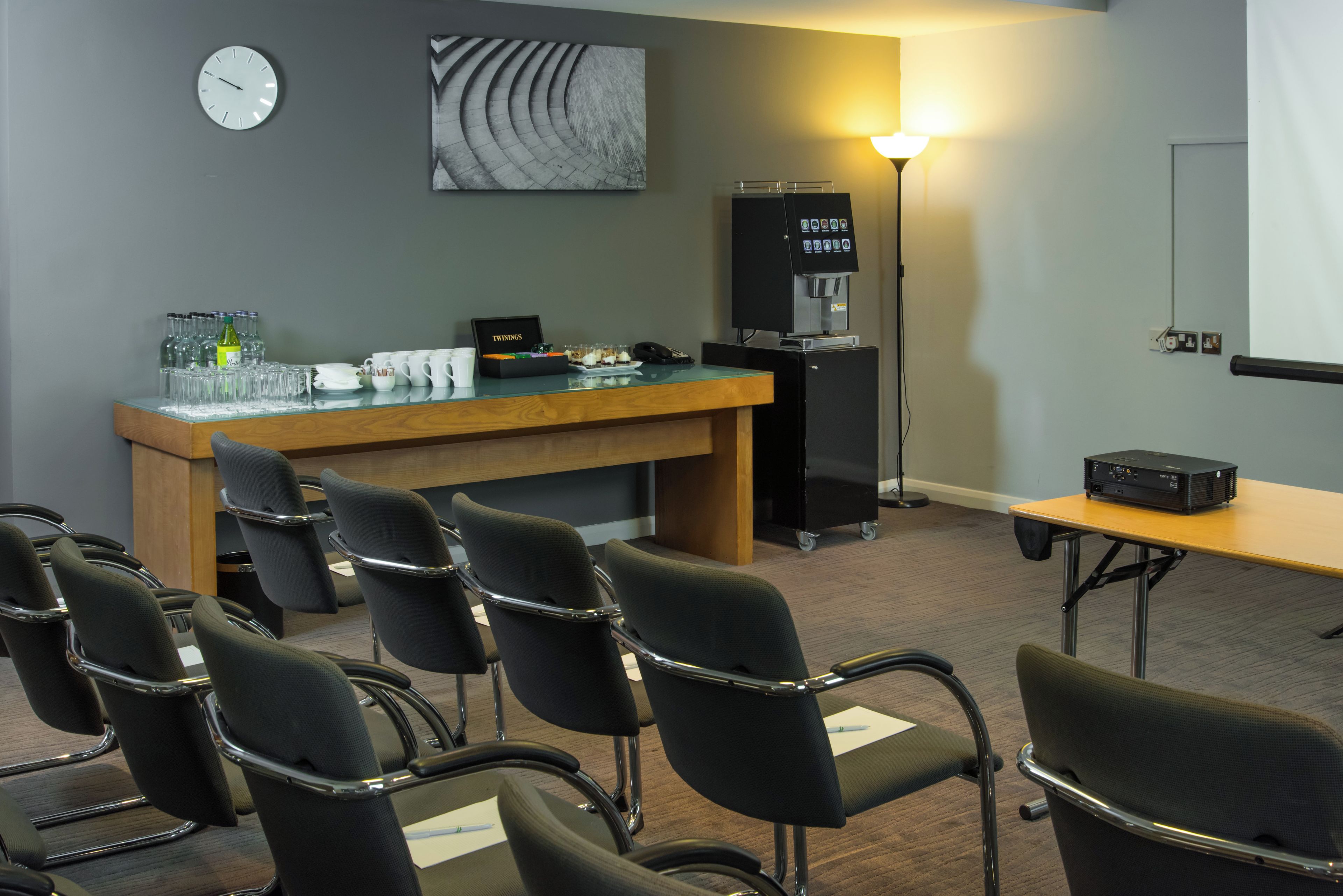Una sala conferenze con un lungo tavolo, diverse sedie, un angolo caffè, un frigorifero, una sveglia e un proiettore.