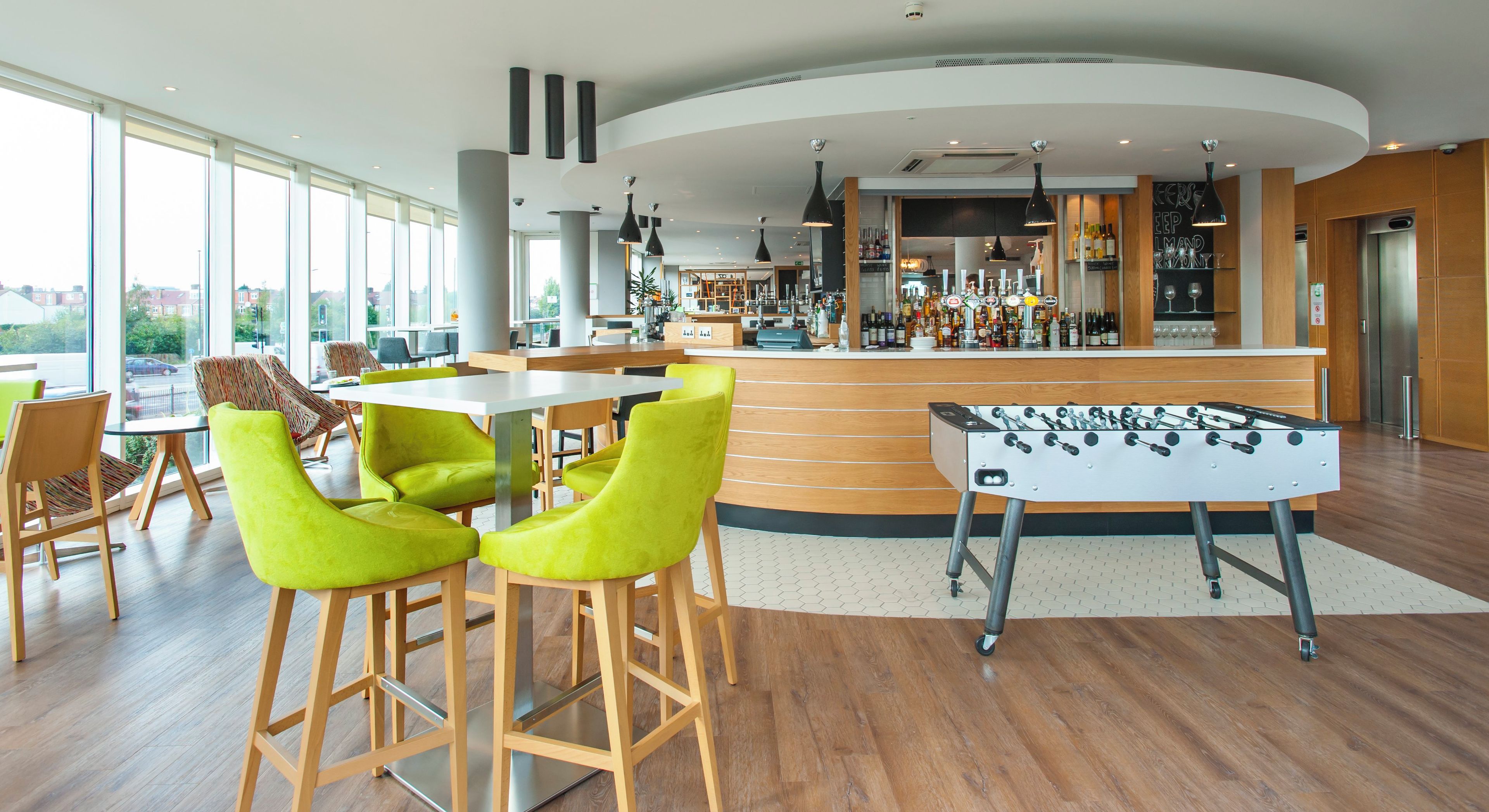 A modern bar area with a foosball table, seating with bright green chairs, and a bar stocked with various bottles.