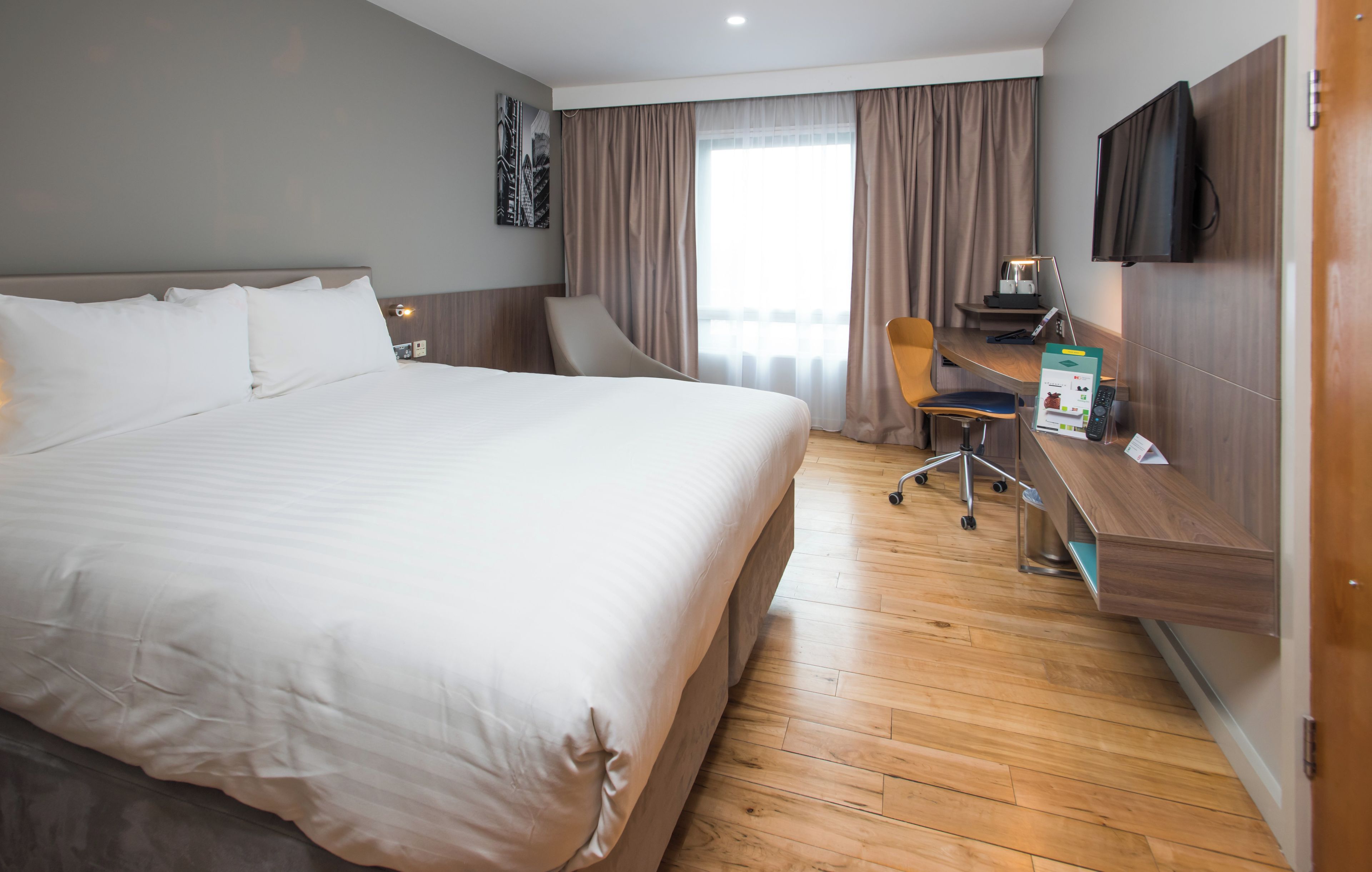 A hotel room with a large bed, a desk with a chair, a flat-screen TV, and a window with curtains.