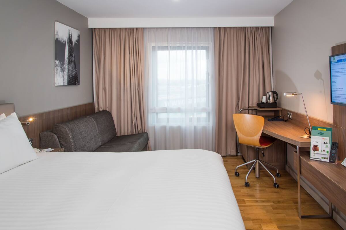 Ein Hotelzimmer mit einem großen Bett, einem Schreibtisch, einem Sessel, einer Lampe und einem Computermonitor.