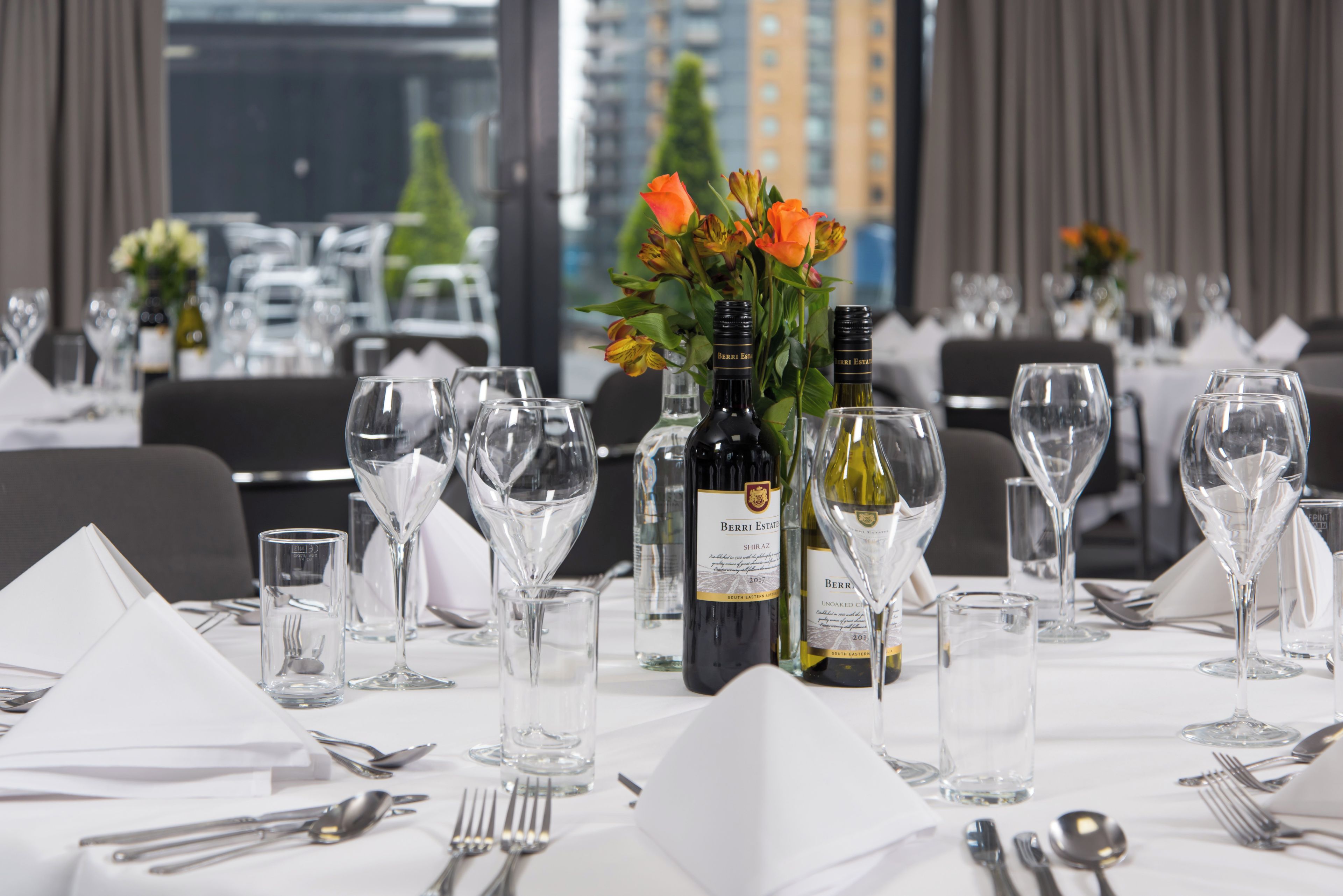 A dining table set with wine glasses, bottles, and cutlery, with a view of buildings in the background.