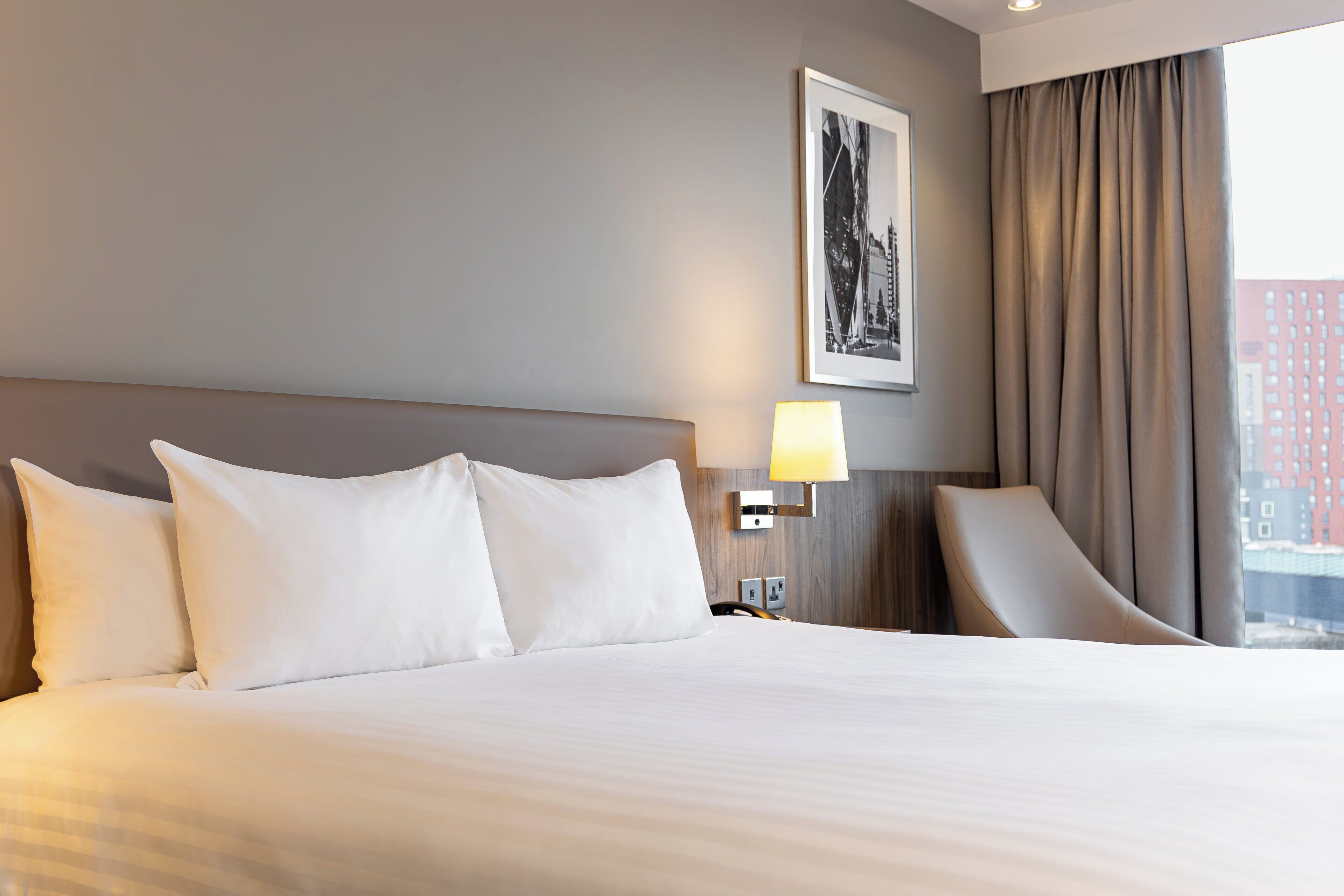 A hotel room with a large bed, a nightstand with a lamp, a chair, and a framed picture on the wall.