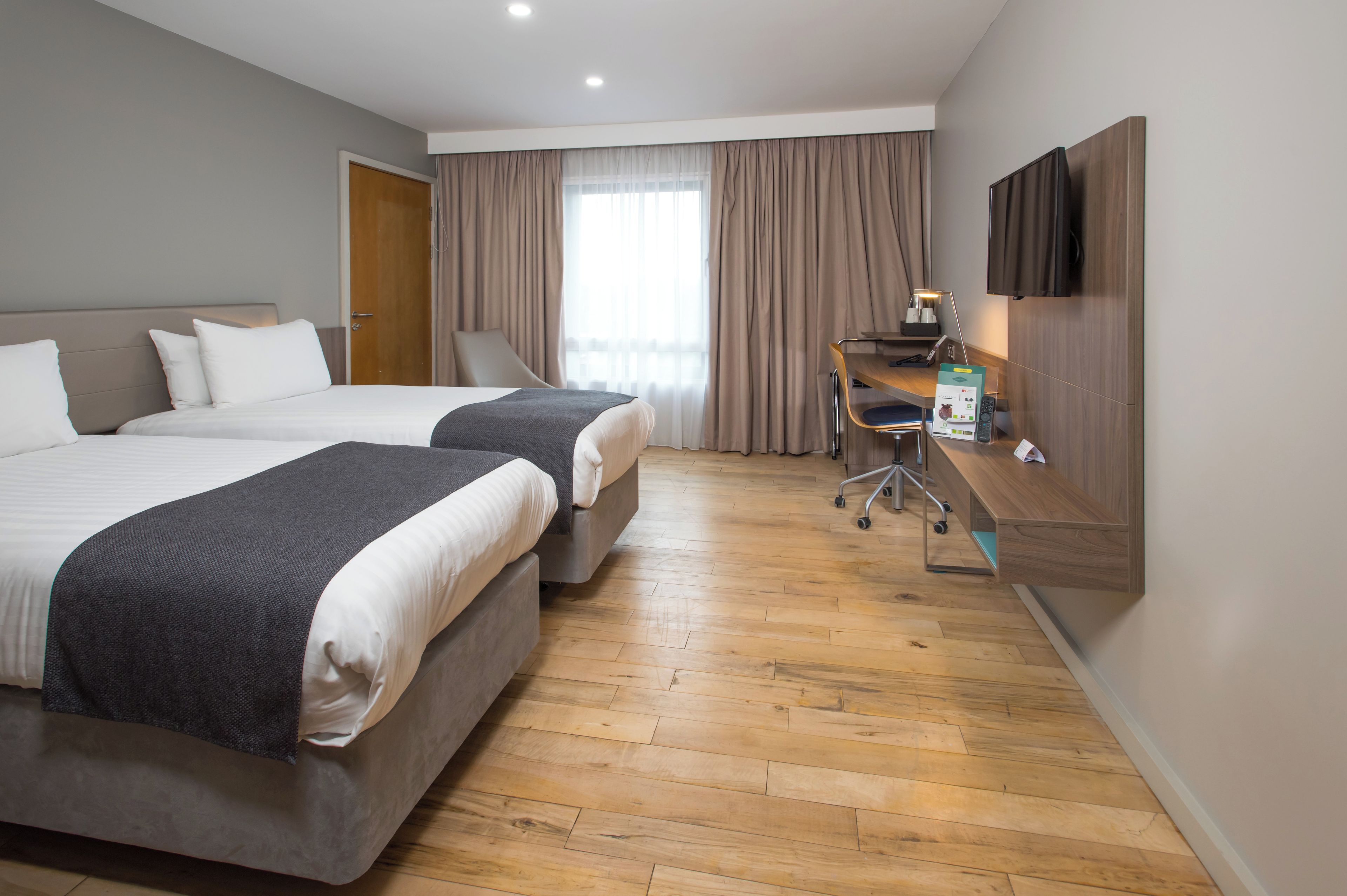 Habitació d'hotel amb dos llits, una escriptori, una cadira, un televisor i una finestra amb cortines.