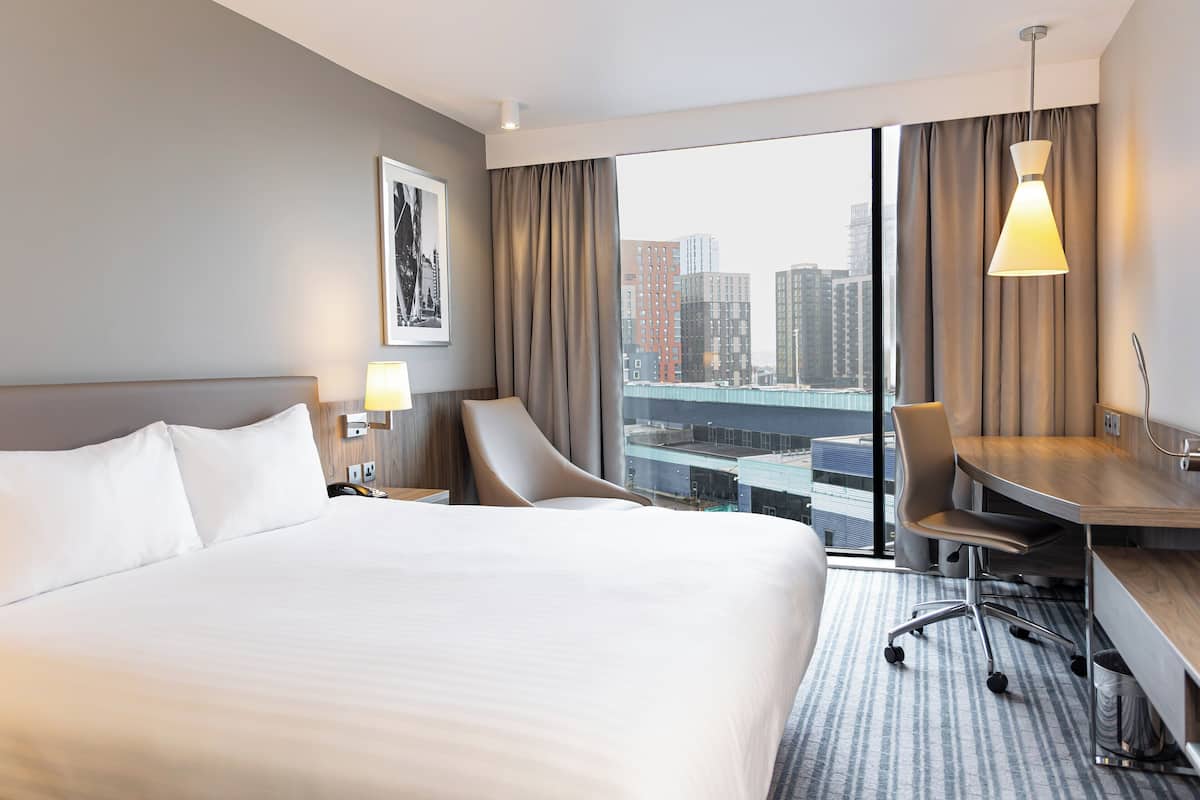 Ein Hotelzimmer mit einem großen Bett, einem Schreibtisch, einem Sessel und Blick auf die Stadt.