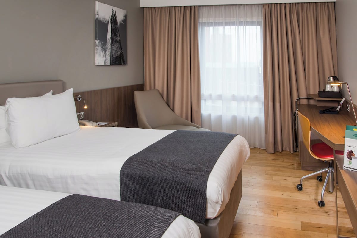 Ein Hotelzimmer mit zwei Betten, einem Schreibtisch mit Stuhl, einer Lampe und einem Fenster mit Vorhängen.
