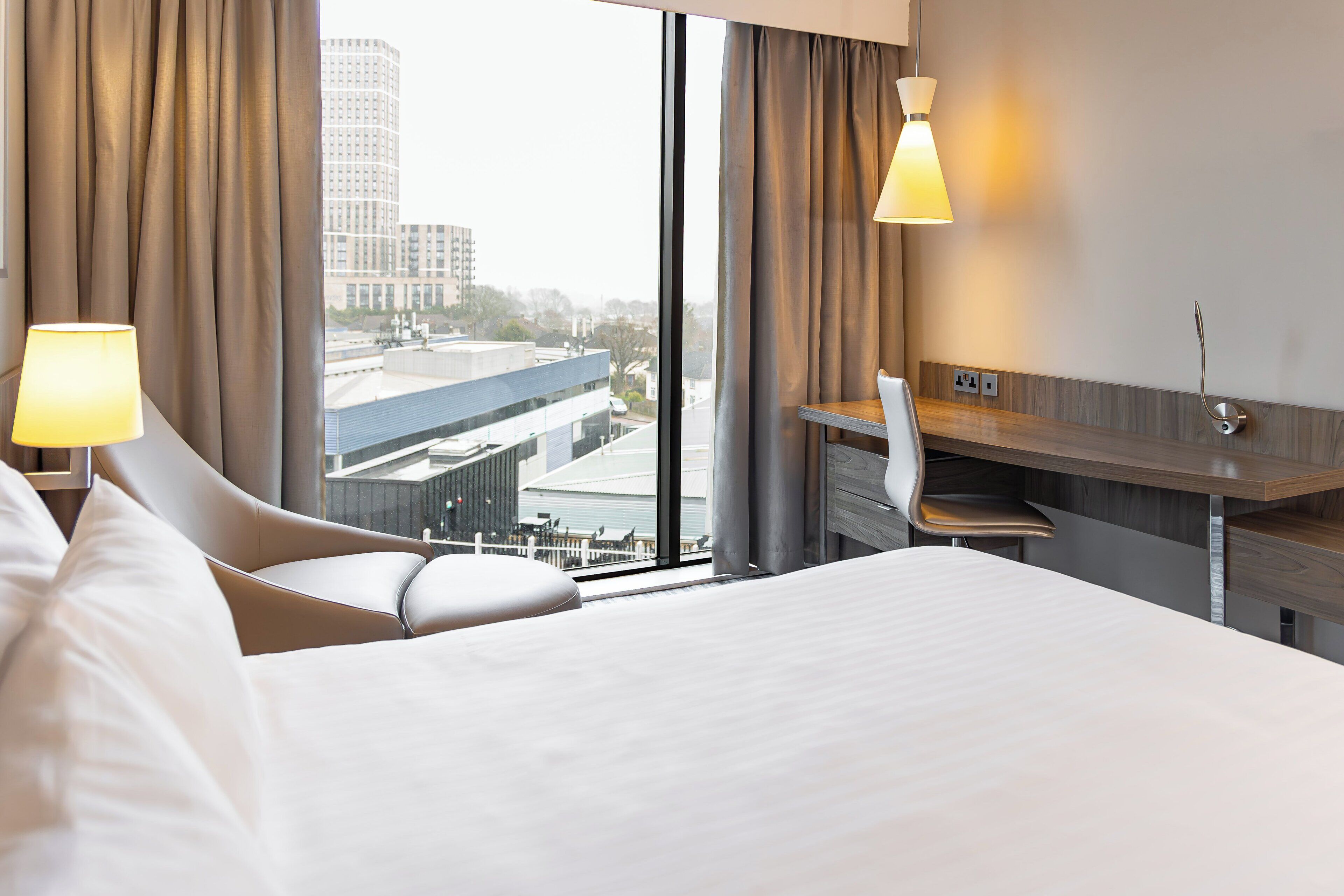 Habitación de hotel con una cama grande, un escritorio y una silla. Hay cortinas y una ventana con vistas a edificios.