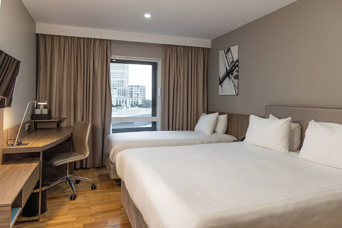 Ein Hotelzimmer mit zwei Betten, einem Schreibtisch, einem Stuhl und einem großen Fenster mit Vorhängen.