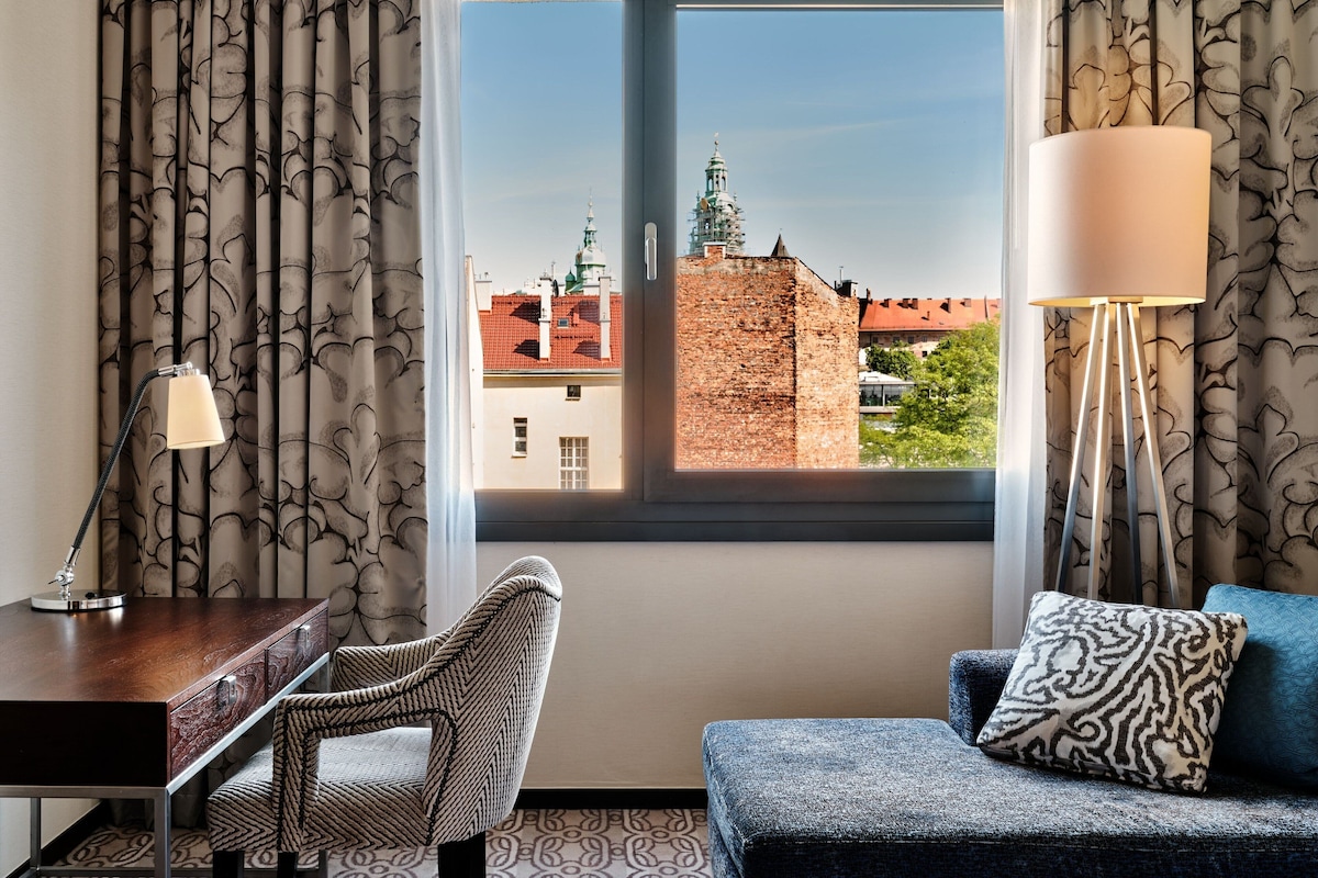 Habitación con vista a un edificio histórico, un escritorio con lámpara, una silla y un sofá con cojines estampados.
