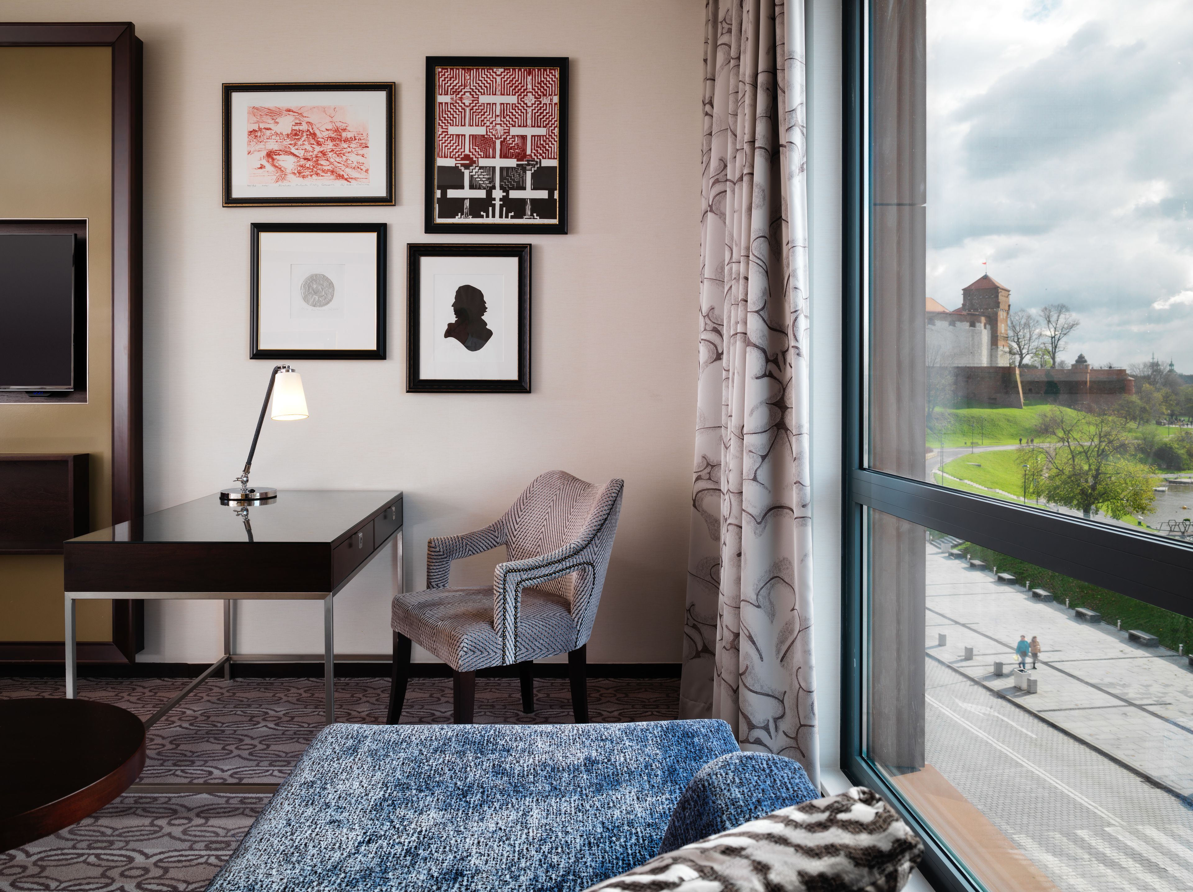 A hotel room with a bed, a desk, a chair, and a large window offering a view of a castle and park.