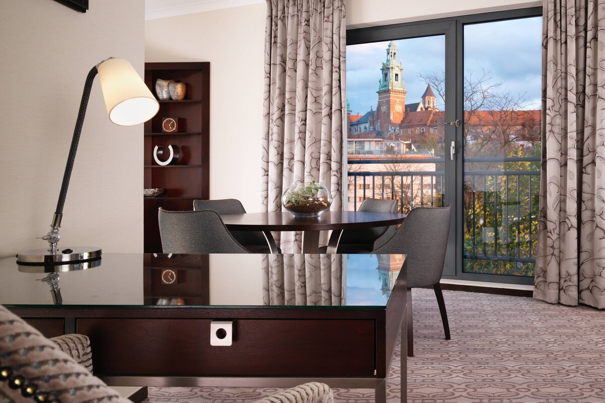 Habitación de hotel con una mesa de comedor, sillas, lámpara, estantería y un ventanal con vistas a un edificio.