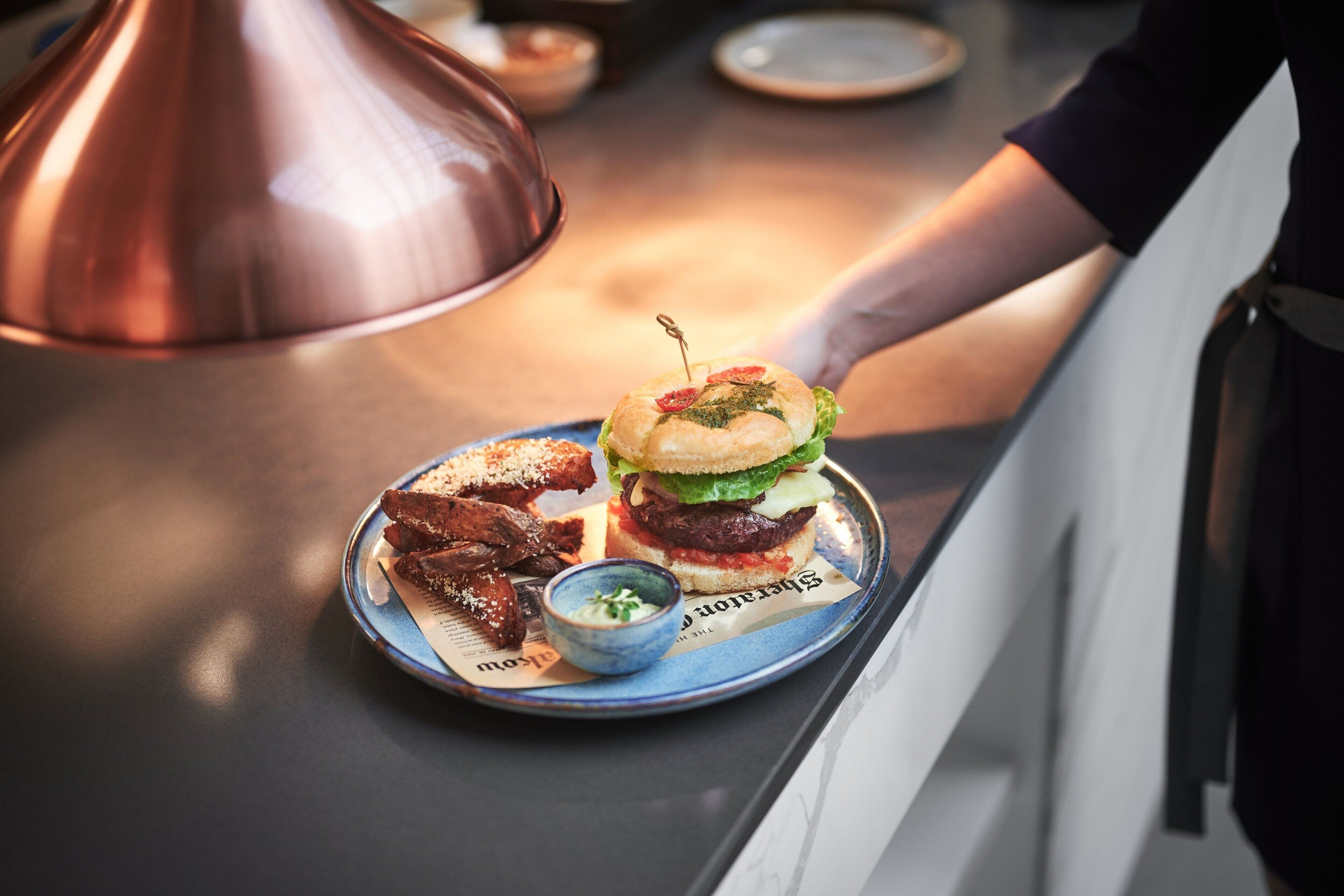 A plate with a burger, fries, and a small bowl of sauce on a table.