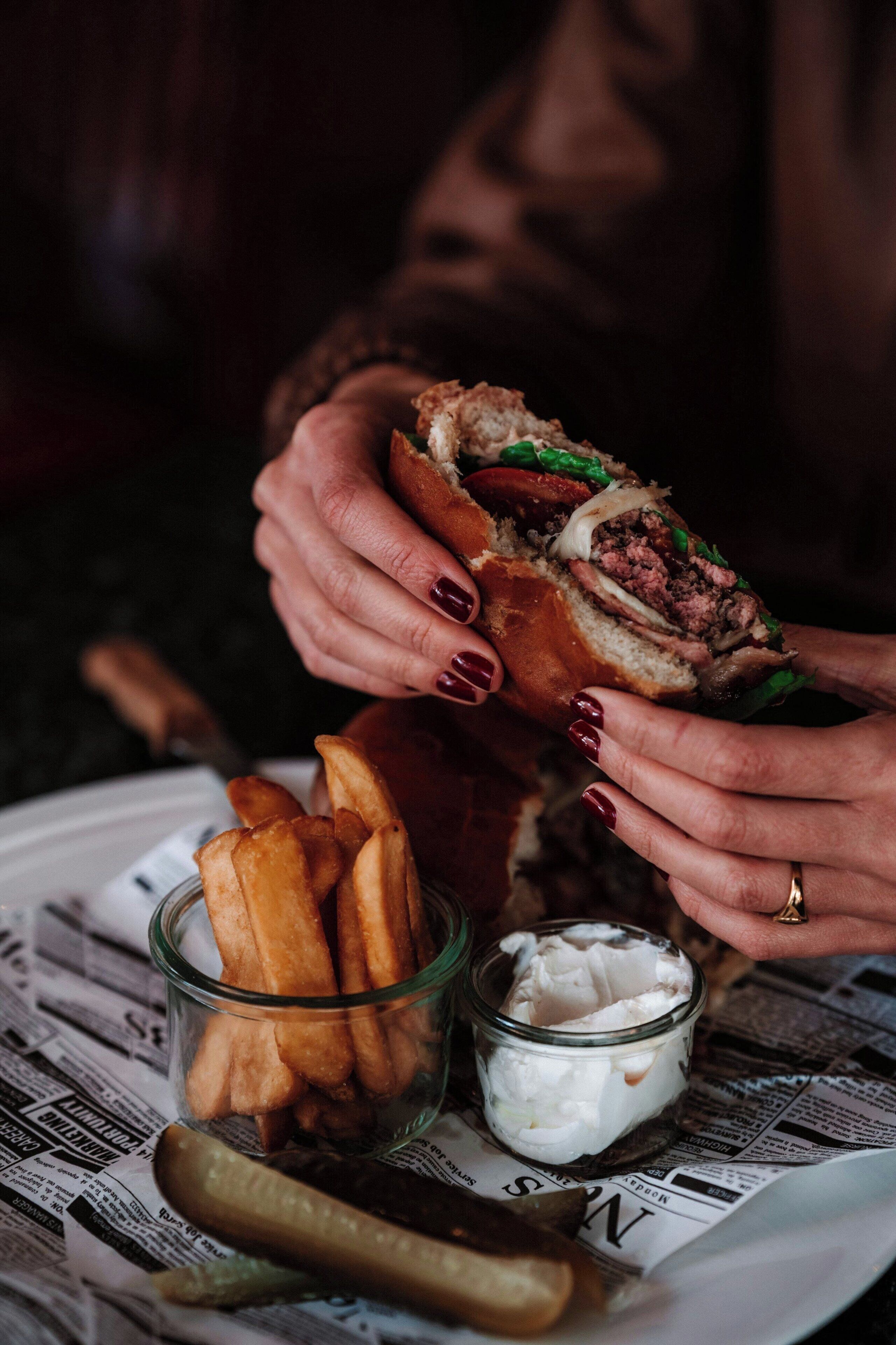 Una persona sosteniendo un sándwich con papas fritas y un recipiente pequeño de salsa sobre una mesa cubierta con un periódico.