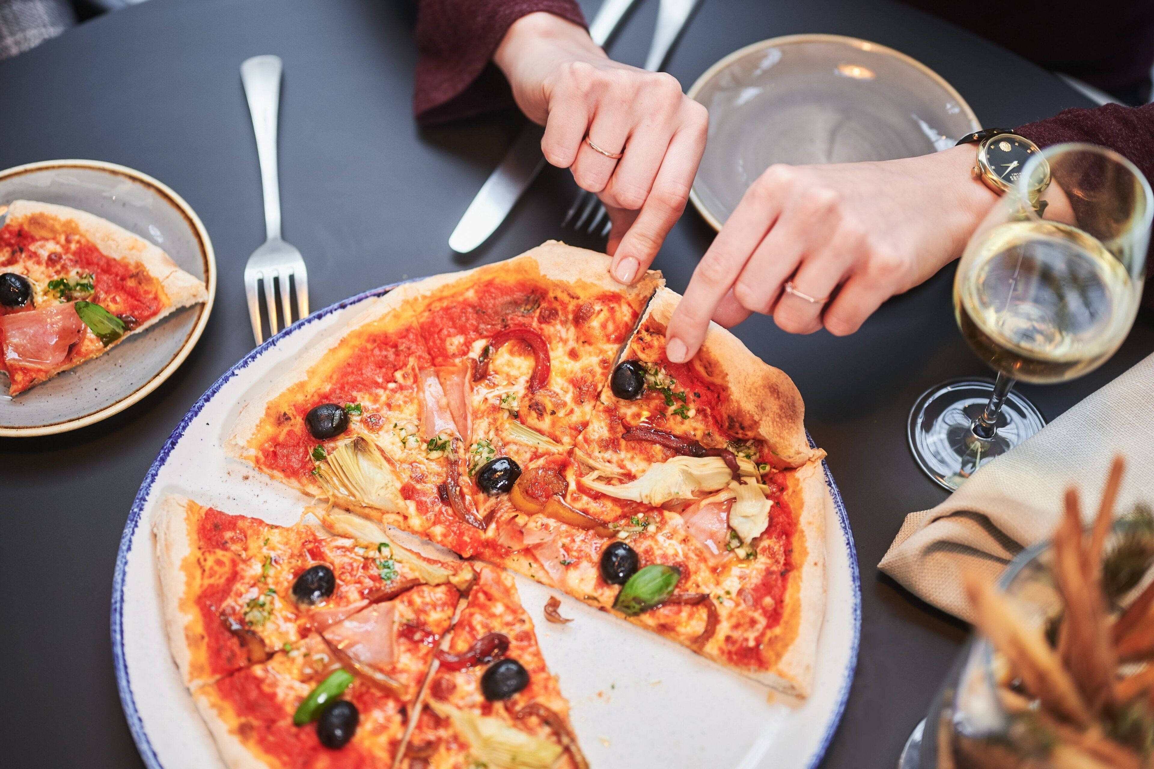 A pizza with various toppings on a plate, a fork, a knife, and a glass of white wine on a table.
