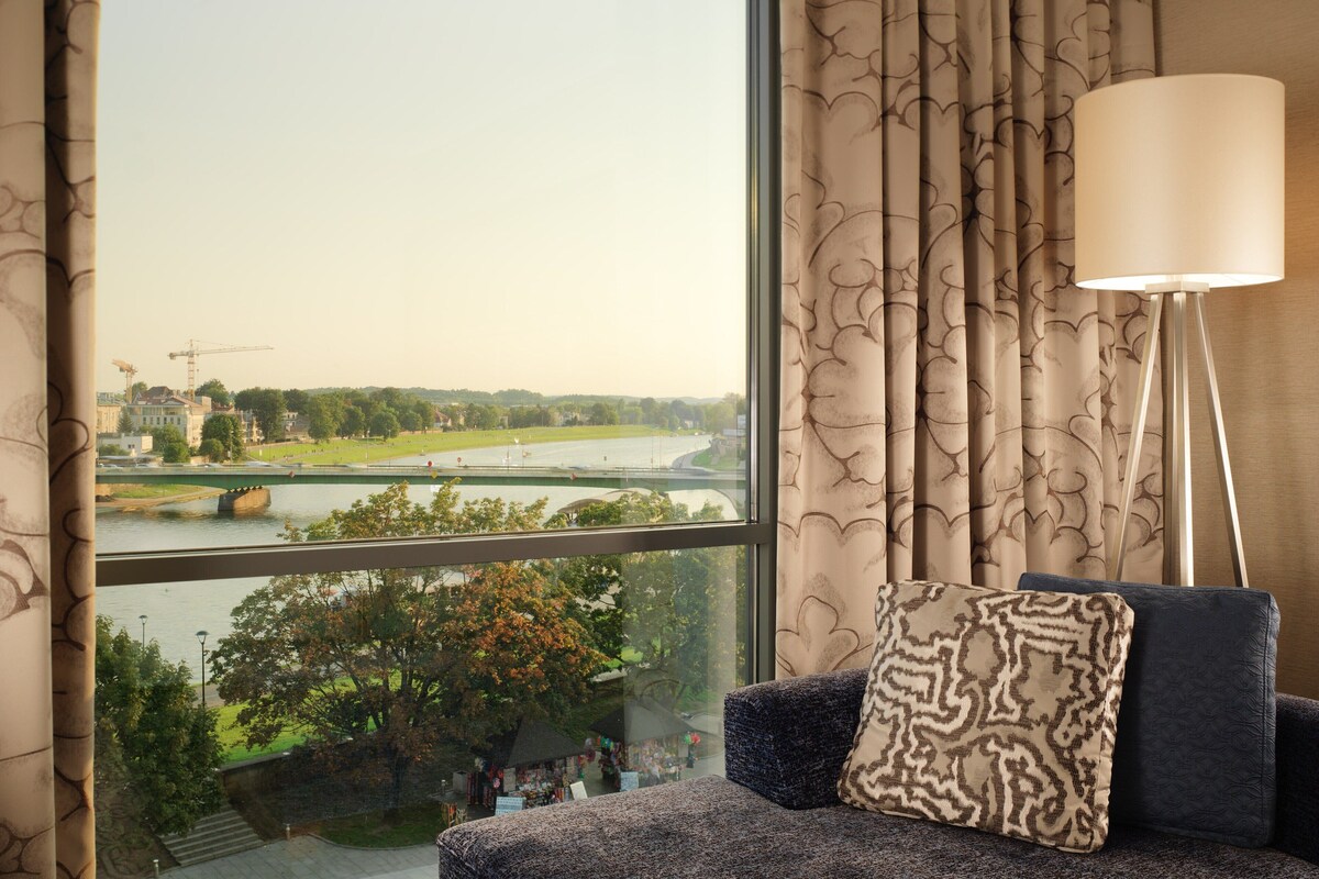 A room with a large window offering a view of a river and a distant cityscape. There is a sofa with patterned cushions and a floor lamp.