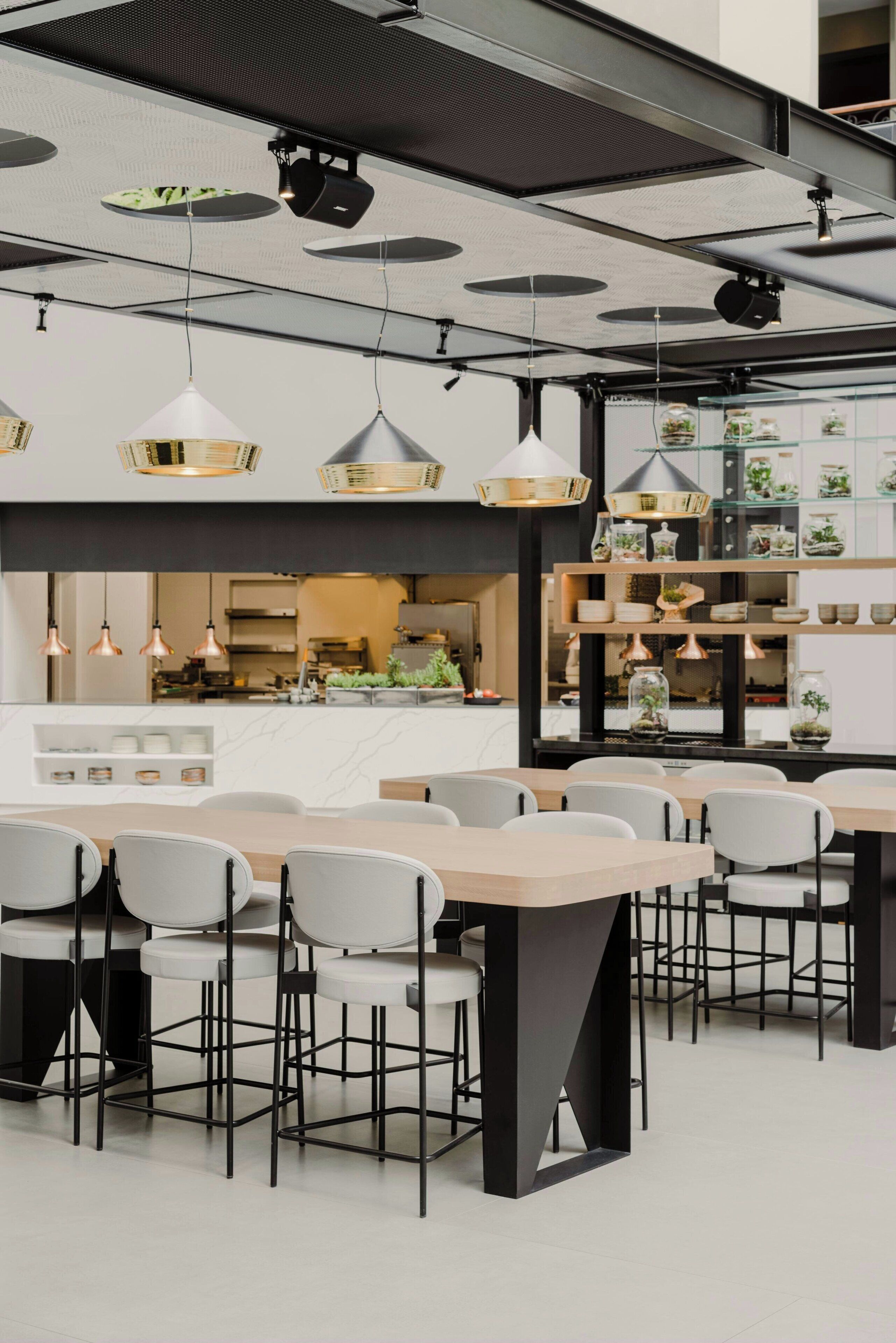 A modern dining area with multiple tables and chairs, a kitchen in the background, and decorative shelves with plants.