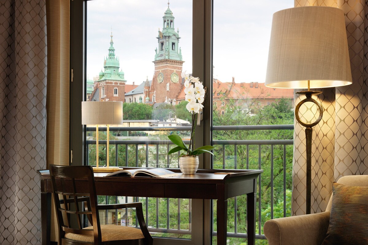A room with a view of a historic building, a table with a book and a potted plant, and a lamp.