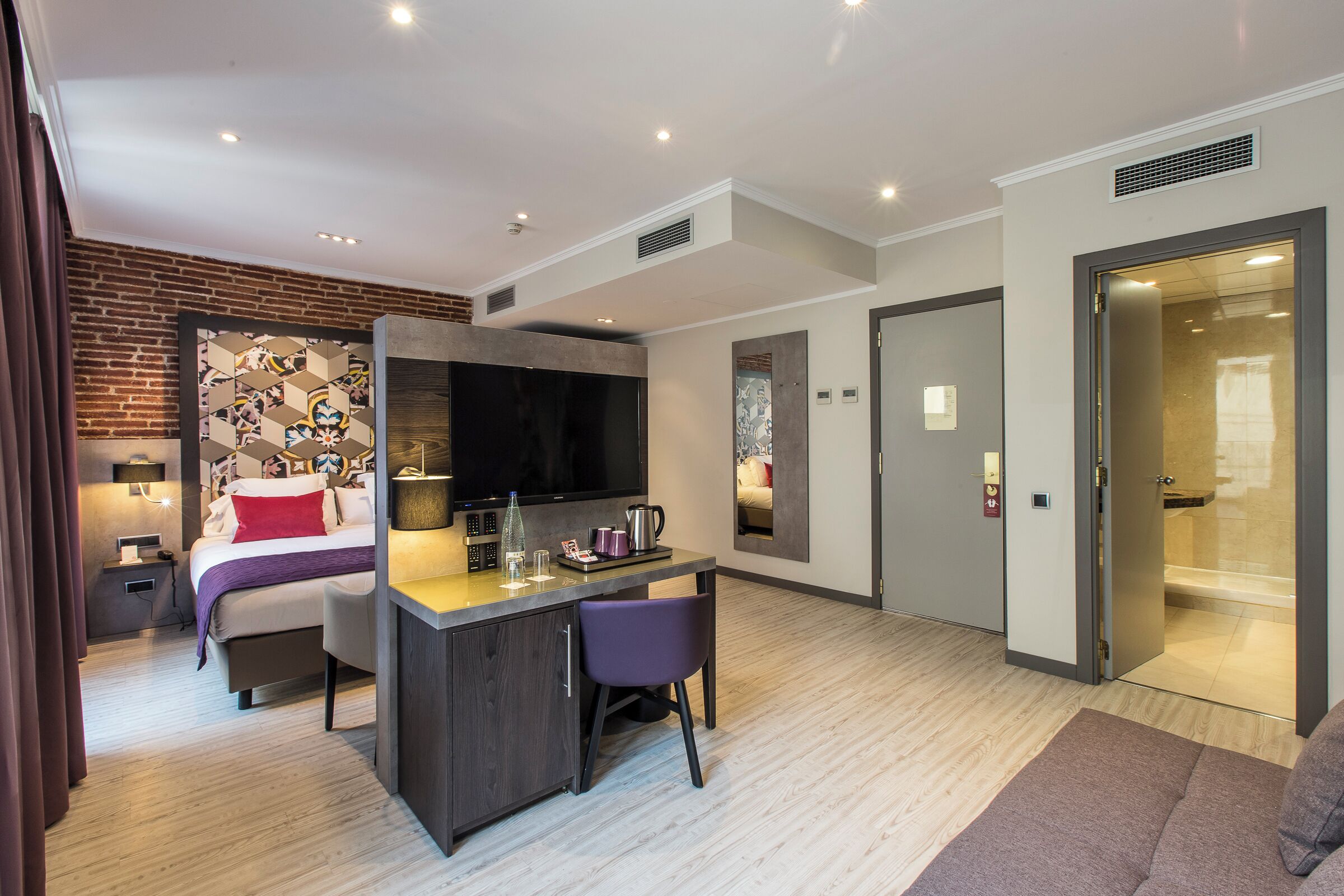 Moderni hotellihuone, jossa on sänky, työpöytä, tuoli, televisio ja suihkutila.